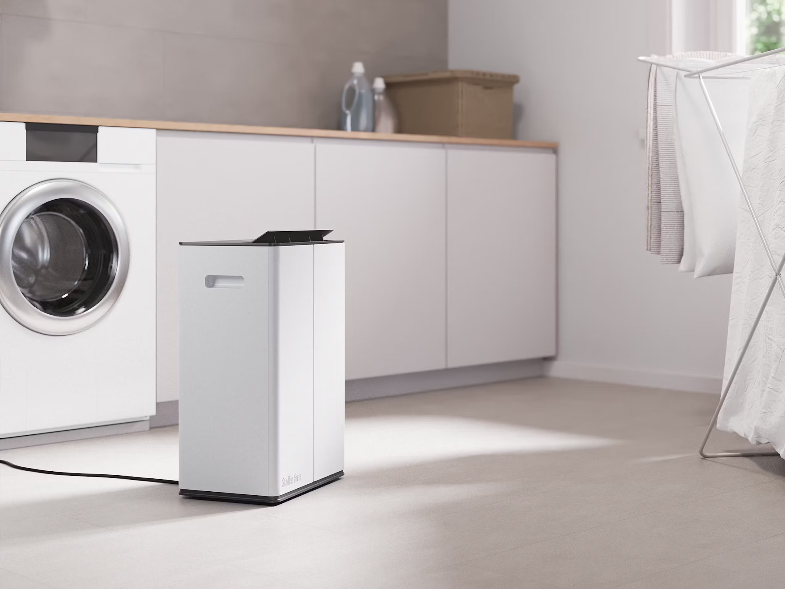Un purificatore d'aria bianco e una lavatrice si trovano in una lavanderia. I vestiti si stanno asciugando su uno stendino.