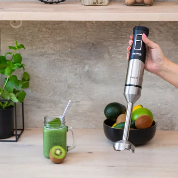 Ręka trzymająca czarno-srebrny blender zanurzeniowy. Zielone smoothie i owoce na stole.