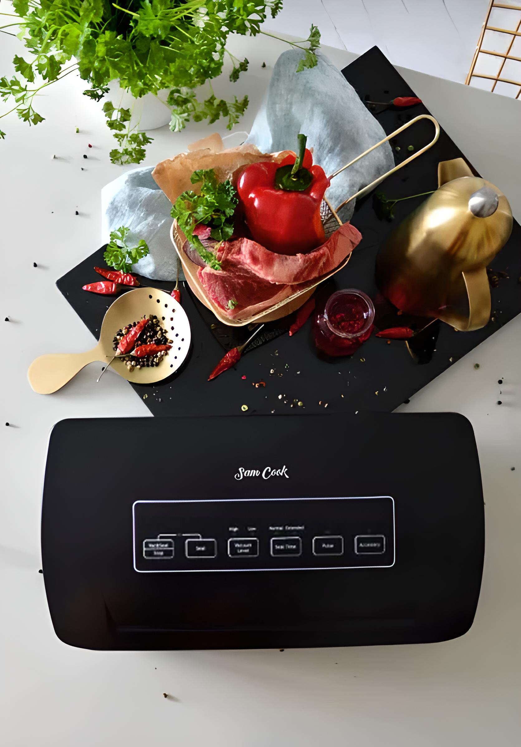 Czarna zgrzewarka próżniowa z jedzeniem, papryką i ziołami na czarnej powierzchni. Biały stół i złoty garnek w tle.