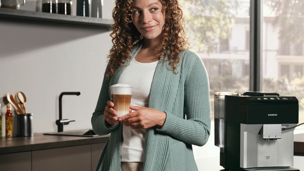 Yeşil bir hırka giymiş bir kadın, bir kahve makinesi olan bir mutfakta latte tutuyor.