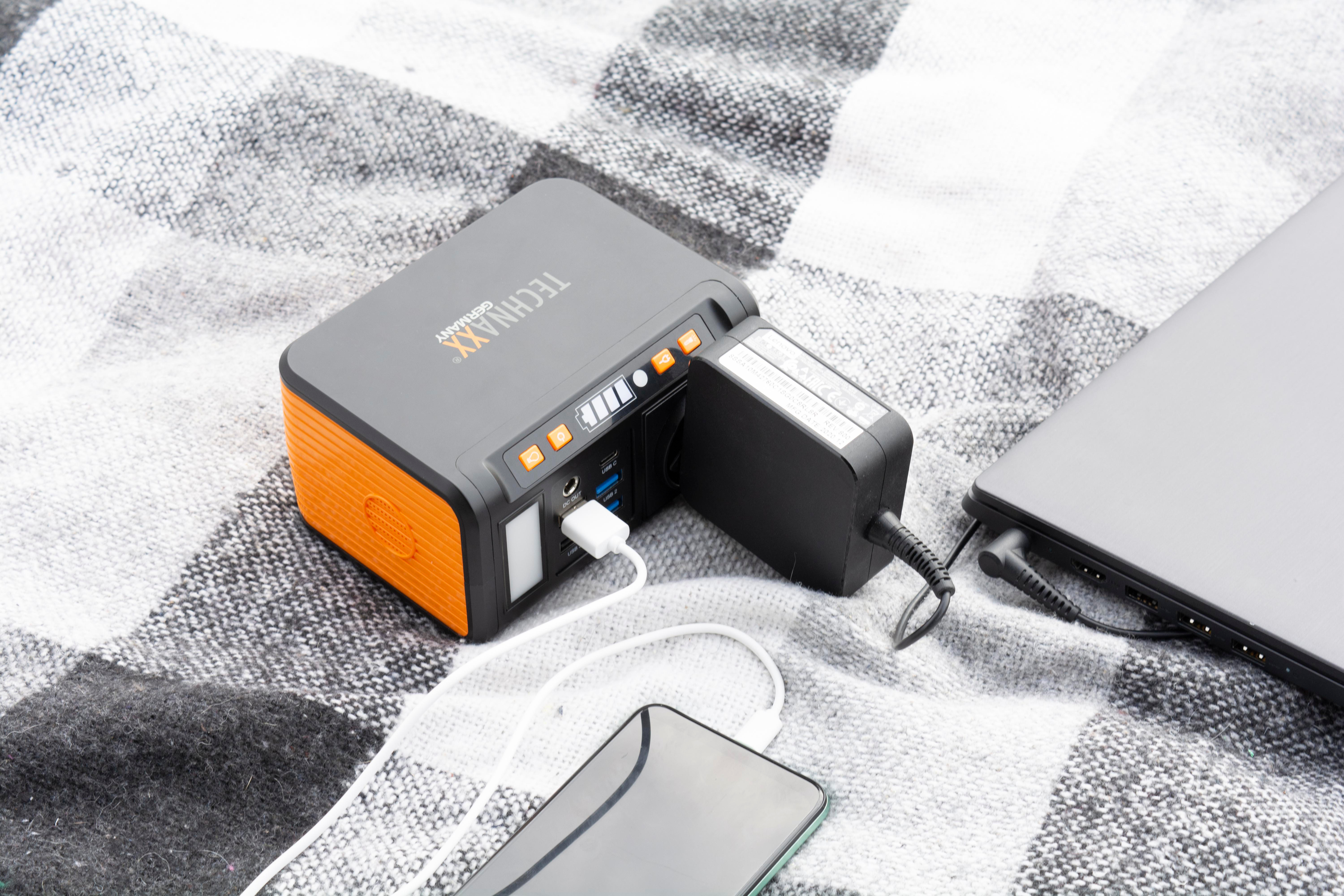 Tragbare Powerstation, lädt Telefon und Laptop. Orange und grau. Karierte Decke im Hintergrund.