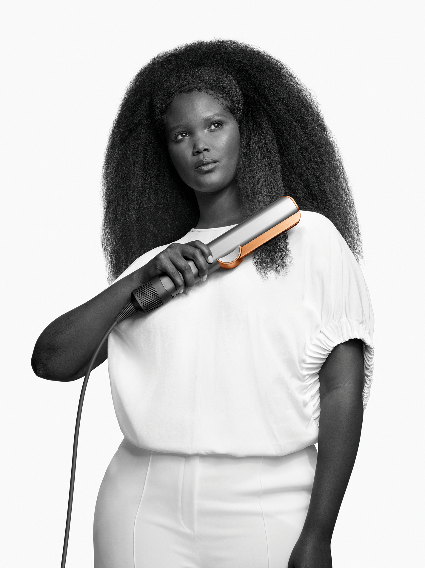 Frau mit Haarglätter in der Hand. Weißes Outfit, schwarze Haare. Hintergrund ist weiß.