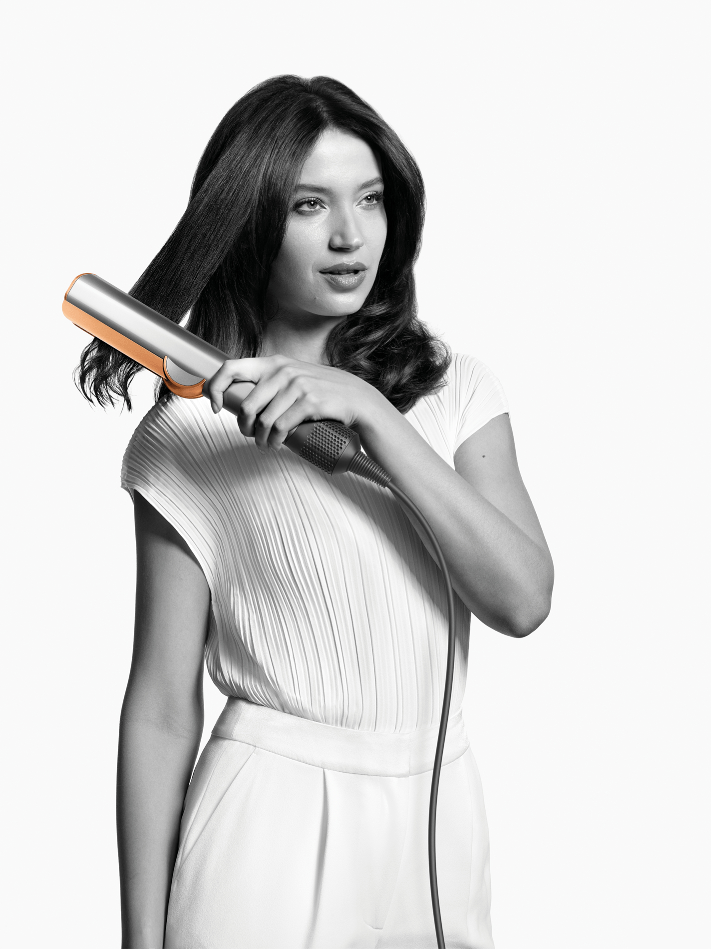 Frau mit glattem Haar, die einen Haarstyler benutzt. Sie trägt ein weißes Oberteil und eine Hose.