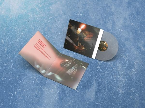 Albumcover mit grauer Schallplatte und offenem Buch mit Kunst auf blauer Oberfläche.