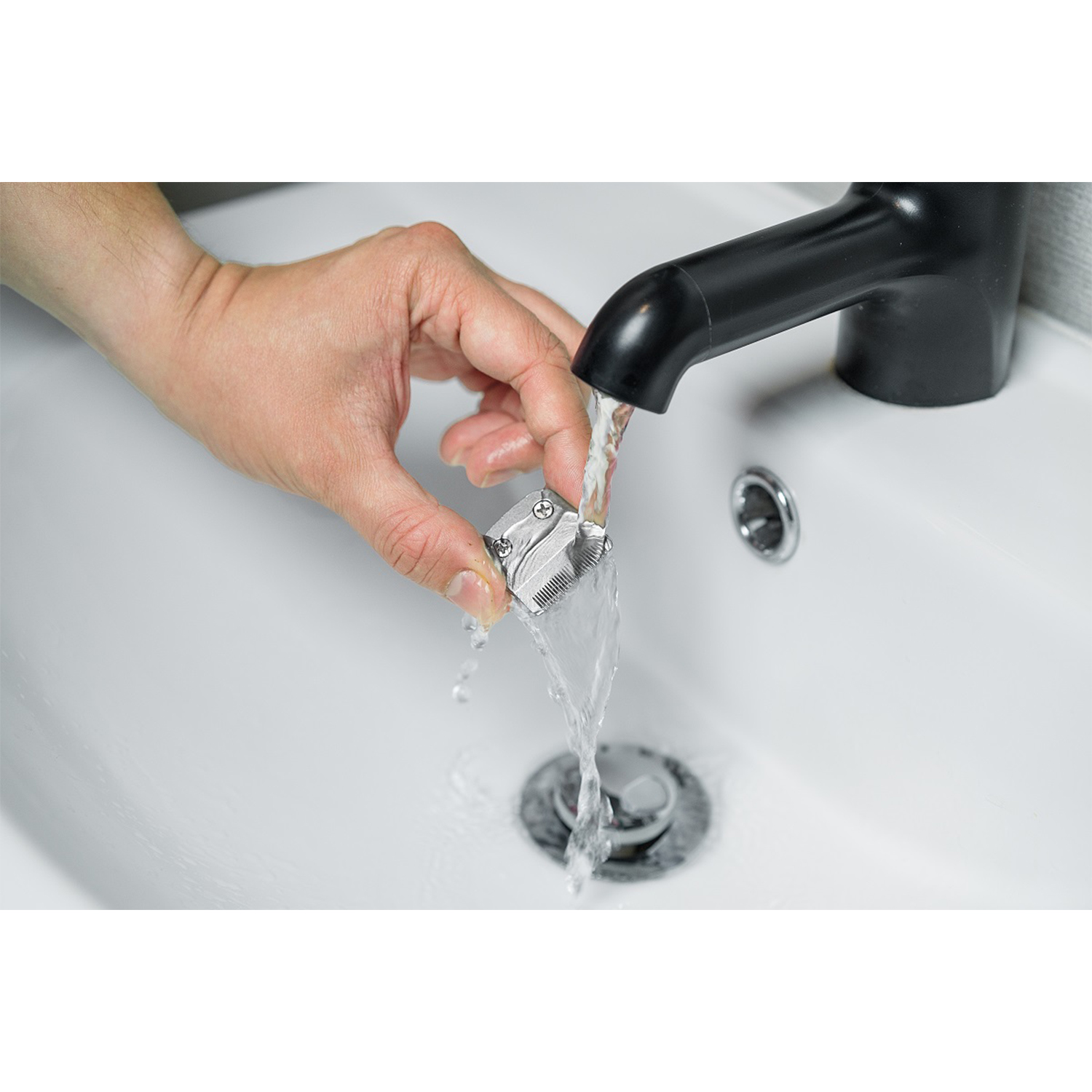 Mano che lava la testina di un trimmer sotto un rubinetto nero. Lavandino bianco. Acqua che scorre.