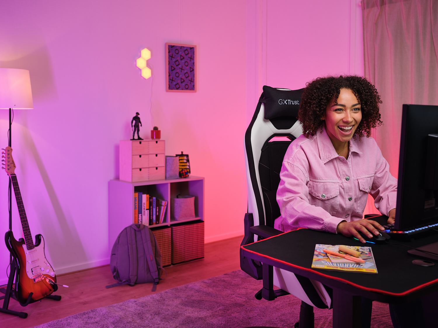 Una donna sorridente in rosa siede a una scrivania con PC, in una stanza con illuminazione rosa e una chitarra a sinistra.
