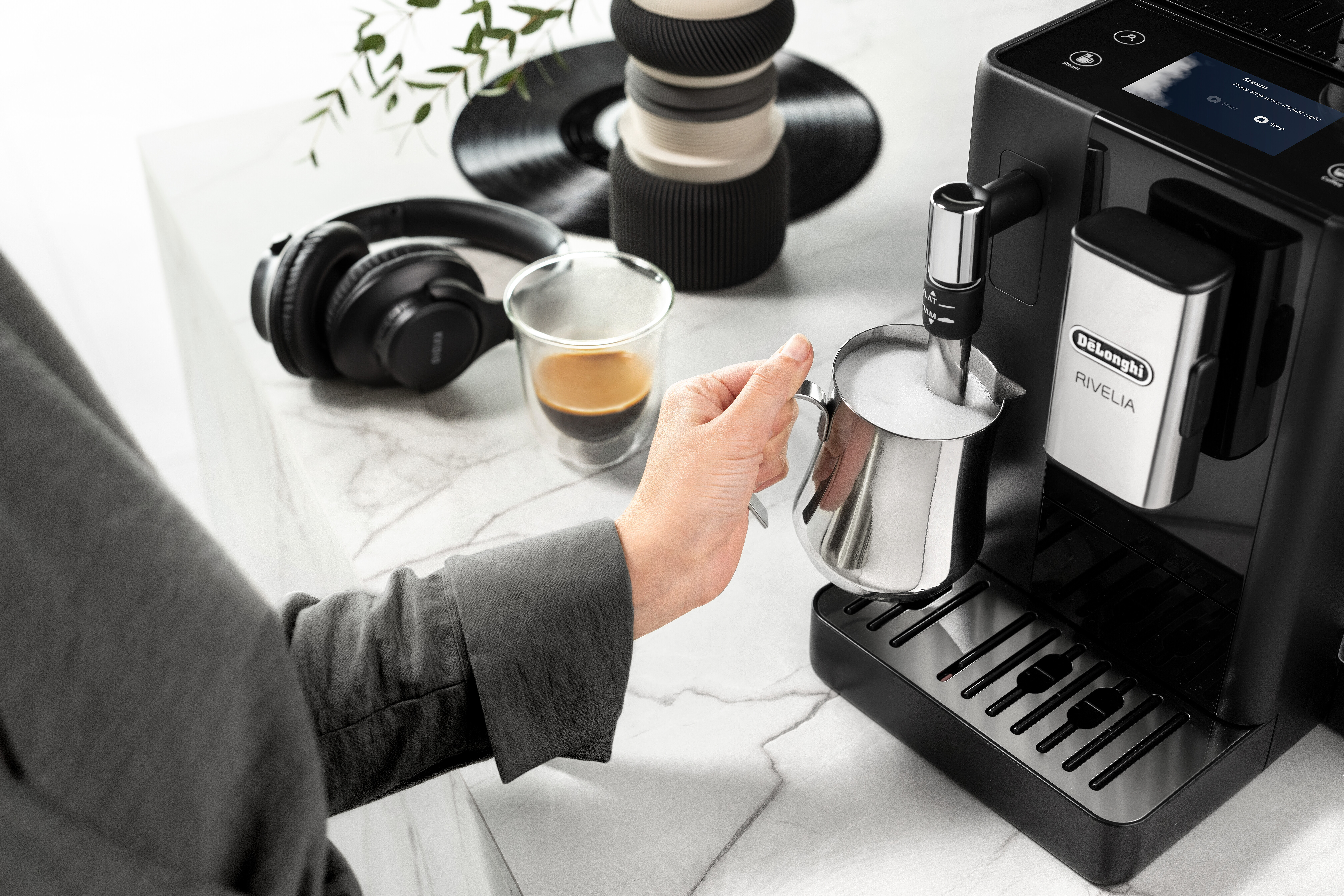 Person gießt Milch aus einem Metallkännchen in die Aufschäumdüse einer Kaffeemaschine.