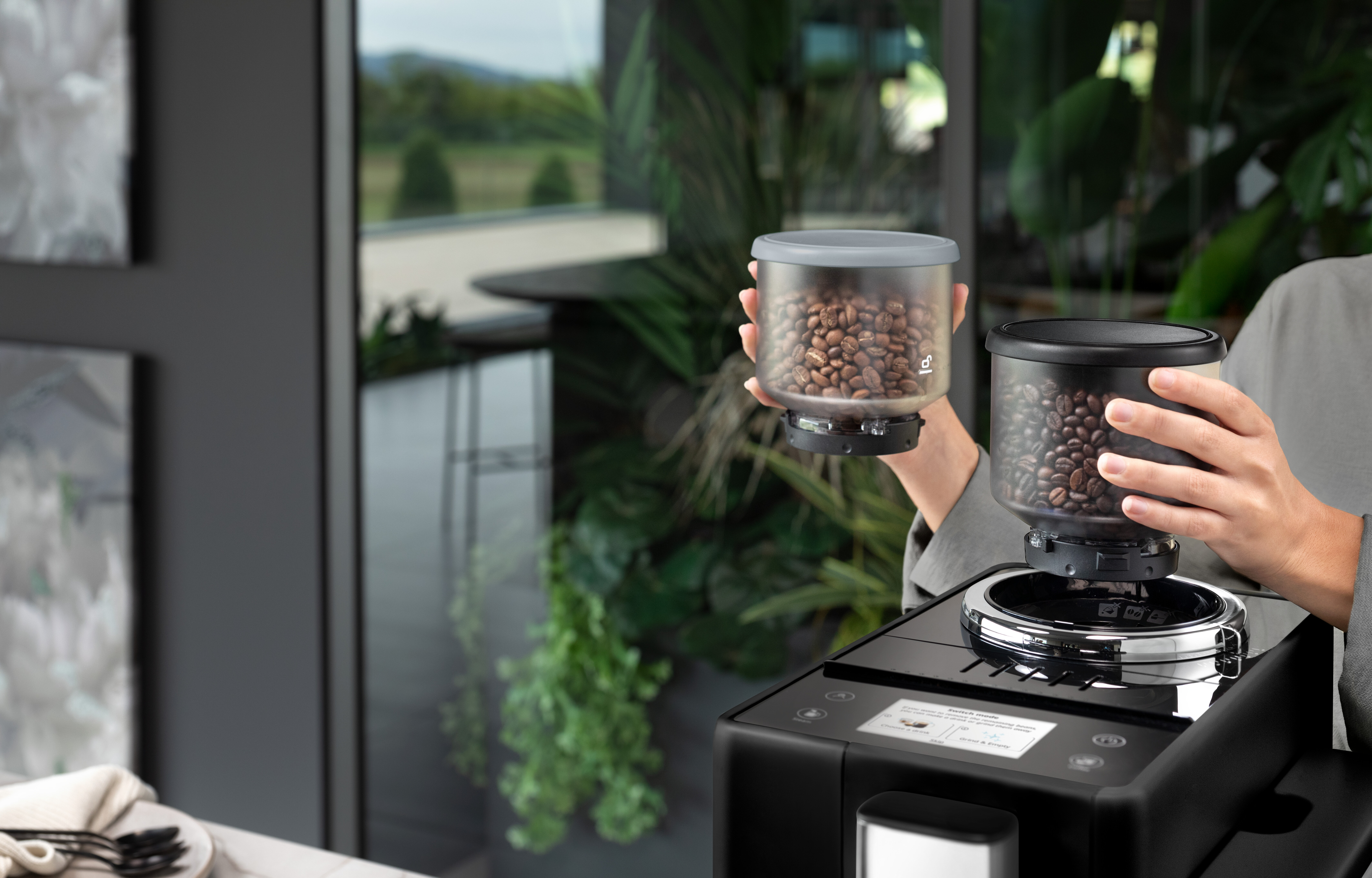 Person hält Behälter mit Kaffeebohnen und gibt sie in eine Kaffeemaschine. Die Maschine ist schwarz.