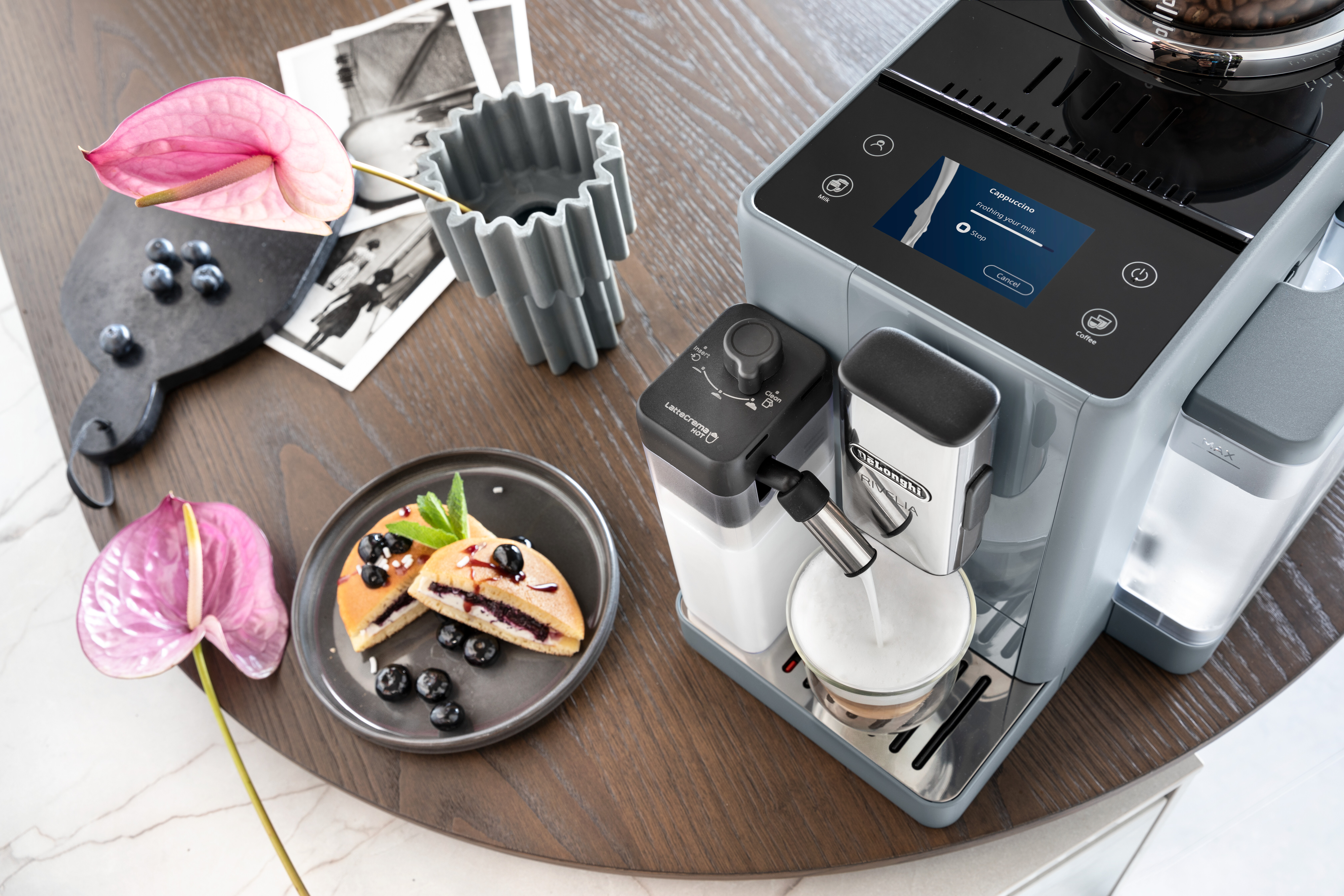 Espressomaschine mit Essen und rosa Blumen auf Holztisch.