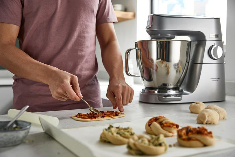 Osoba rozsmarowuje sos na cieście obok miksera stojącego, z przygotowanymi roladkami pizzy.