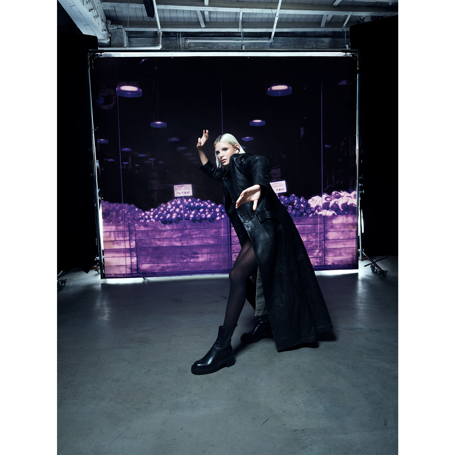 Una donna con un cappotto nero posa in uno studio. Uno sfondo illuminato di viola raffigura una scena di negozio di alimentari.
