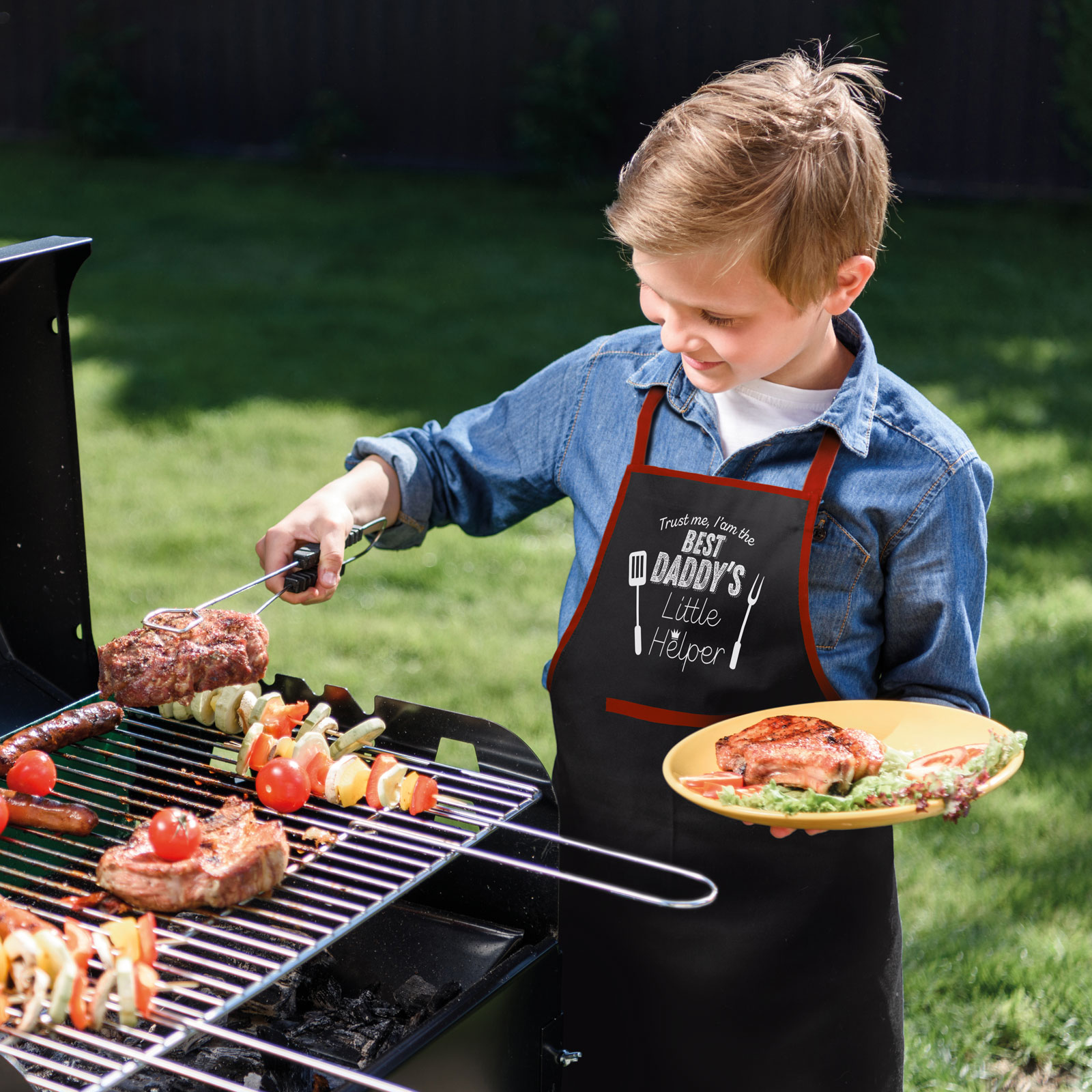 Fiú húst süt grillezőn, a kötényen a felirat: 'Best Daddy's Little Helper', zöld gyepen.