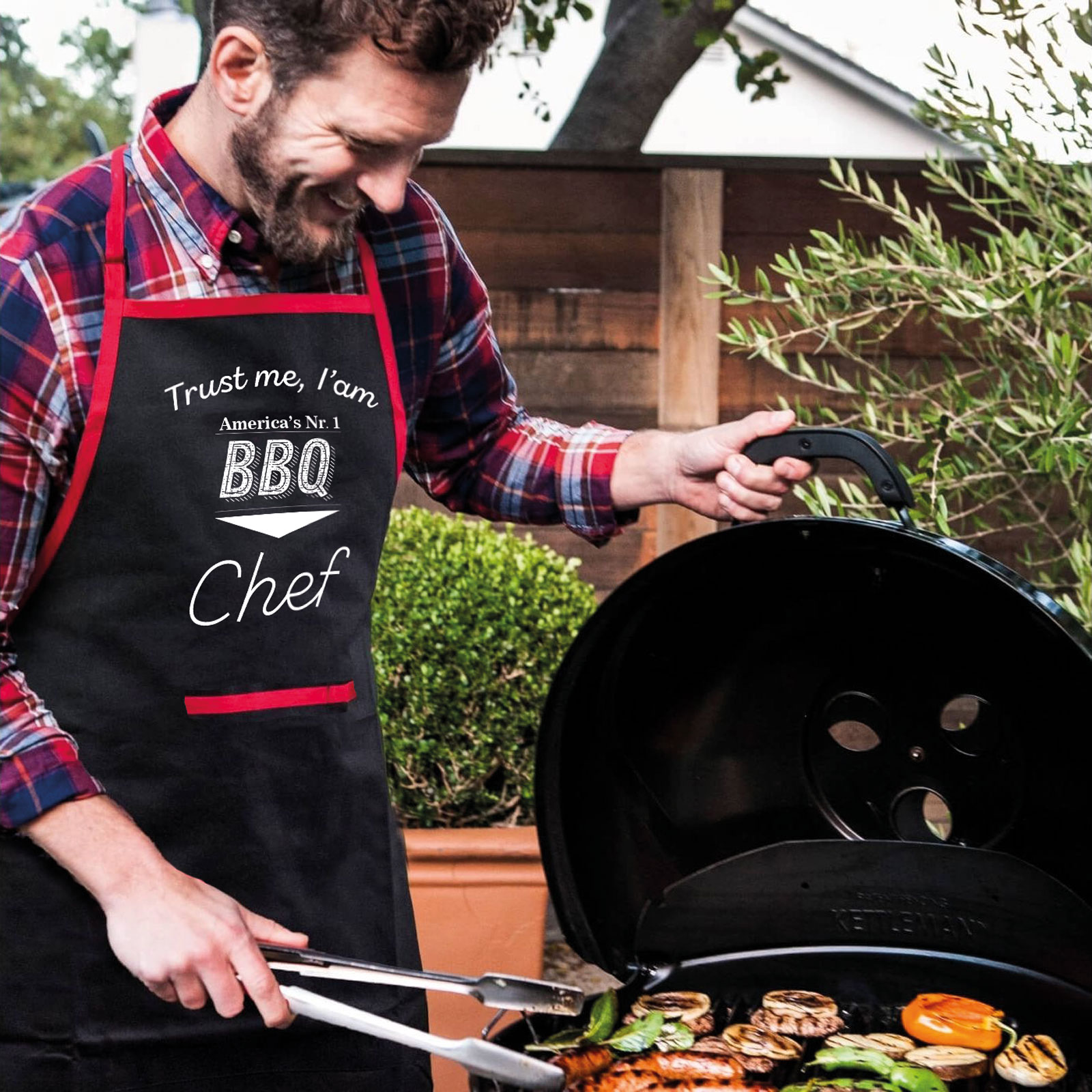 Férfi kötényben, ételt süt, mosolyog, BBQ eszközöket használ.