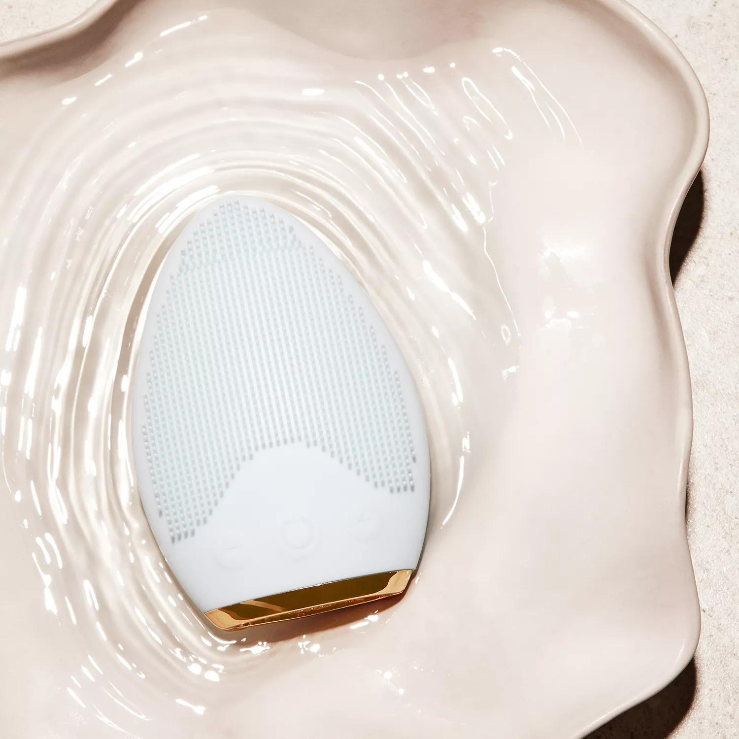 Ein weißes Gesichtsreinigungsgerät in einer weißen, wellenförmigen, muschelförmigen Schale mit Wasser.