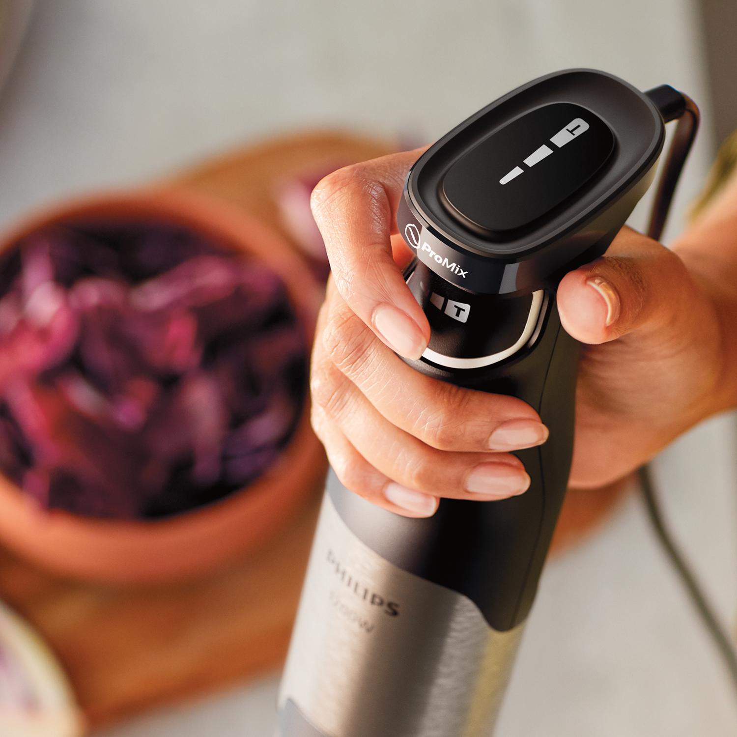 Hand houdt een zwarte Philips staafmixer vast. Een kom met bieten op de achtergrond.