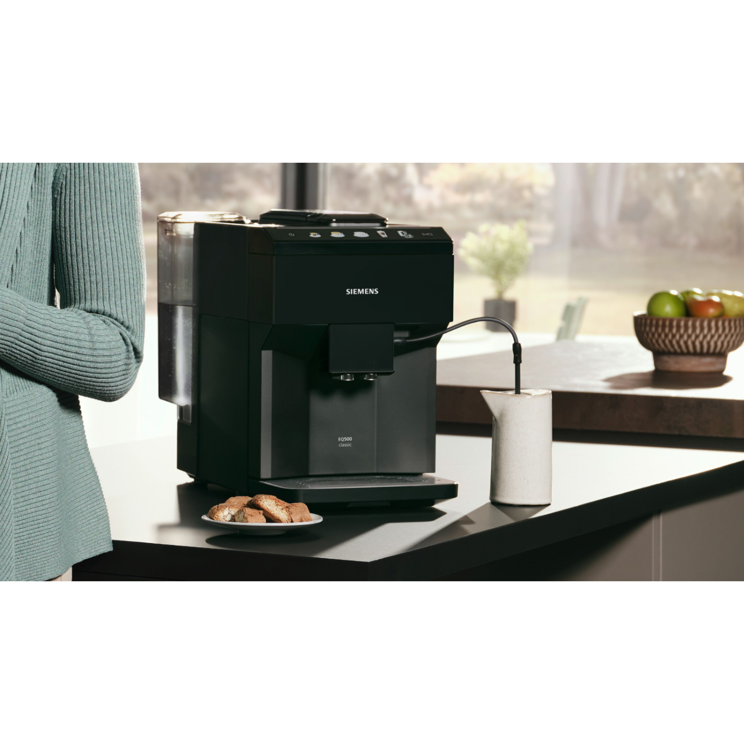 A woman stands near a black coffee machine, with a white carafe and cookies visible.