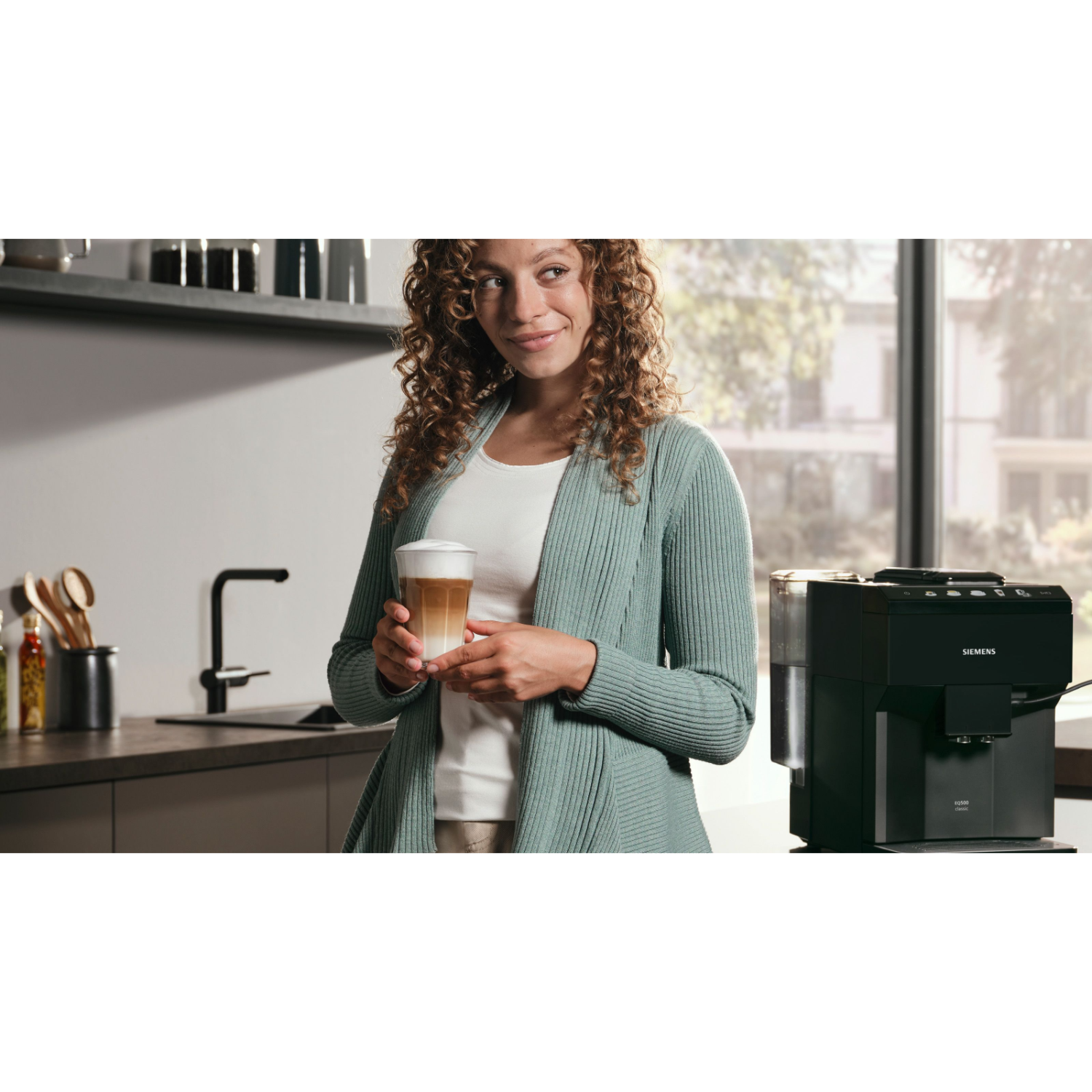 A woman with curly hair holds a latte. A black Siemens coffee machine is on the right.