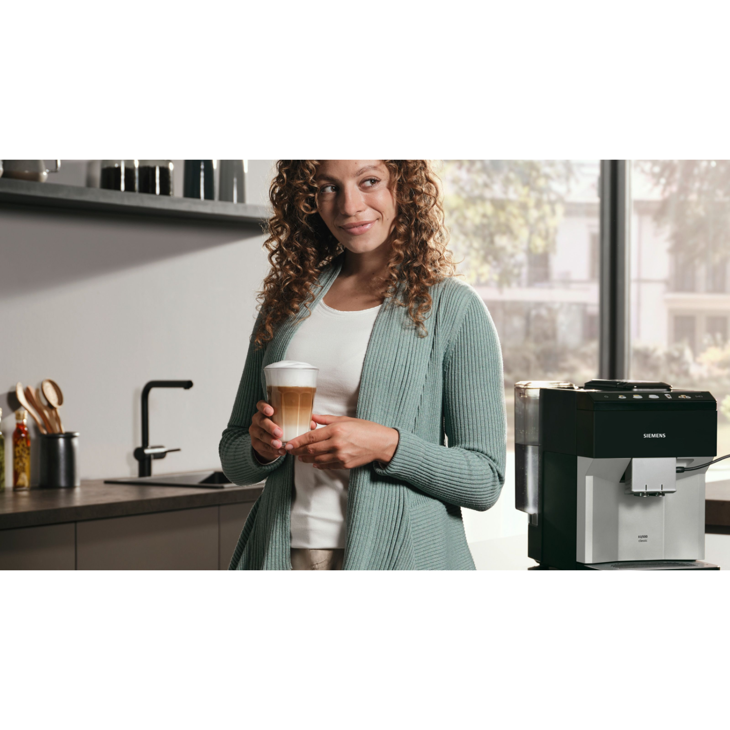 Een vrouw die een latte vasthoudt, staand in een keuken met een Siemens koffiemachine.