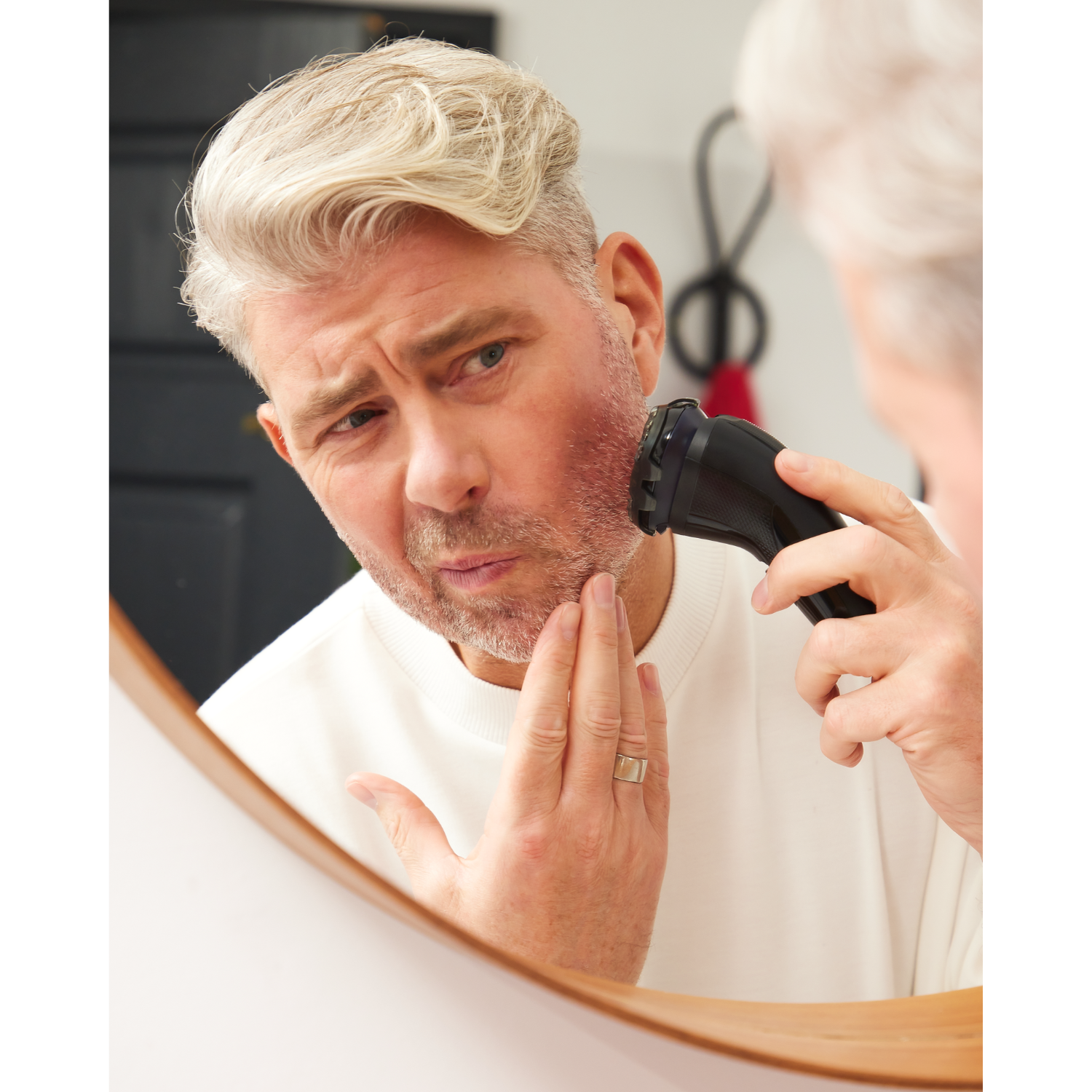 A man shaves his red-irritated face in the mirror with an electric razor.