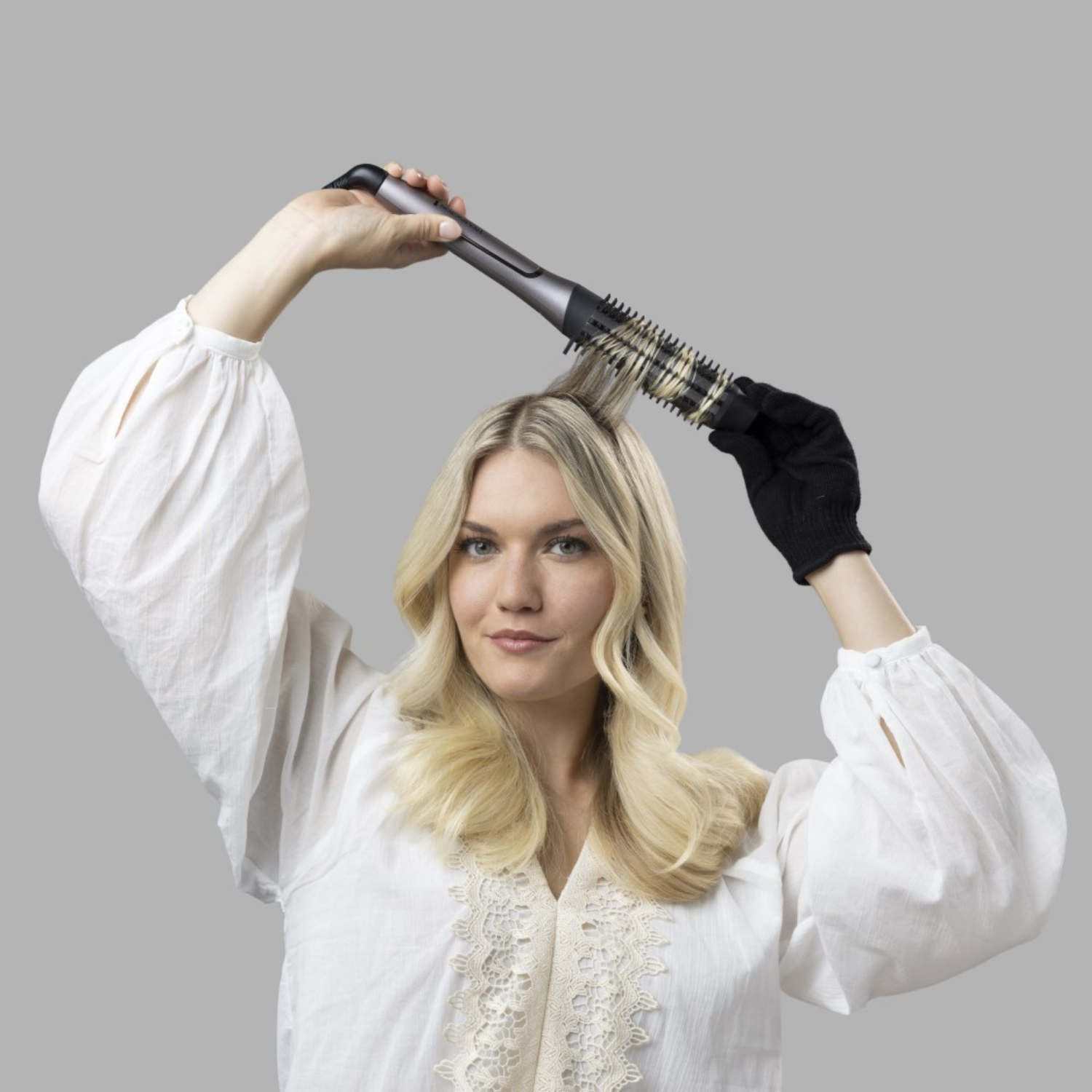 Een vrouw die haar haar stylet met een krulborstel, terwijl ze een wit shirt en zwarte handschoenen draagt.
