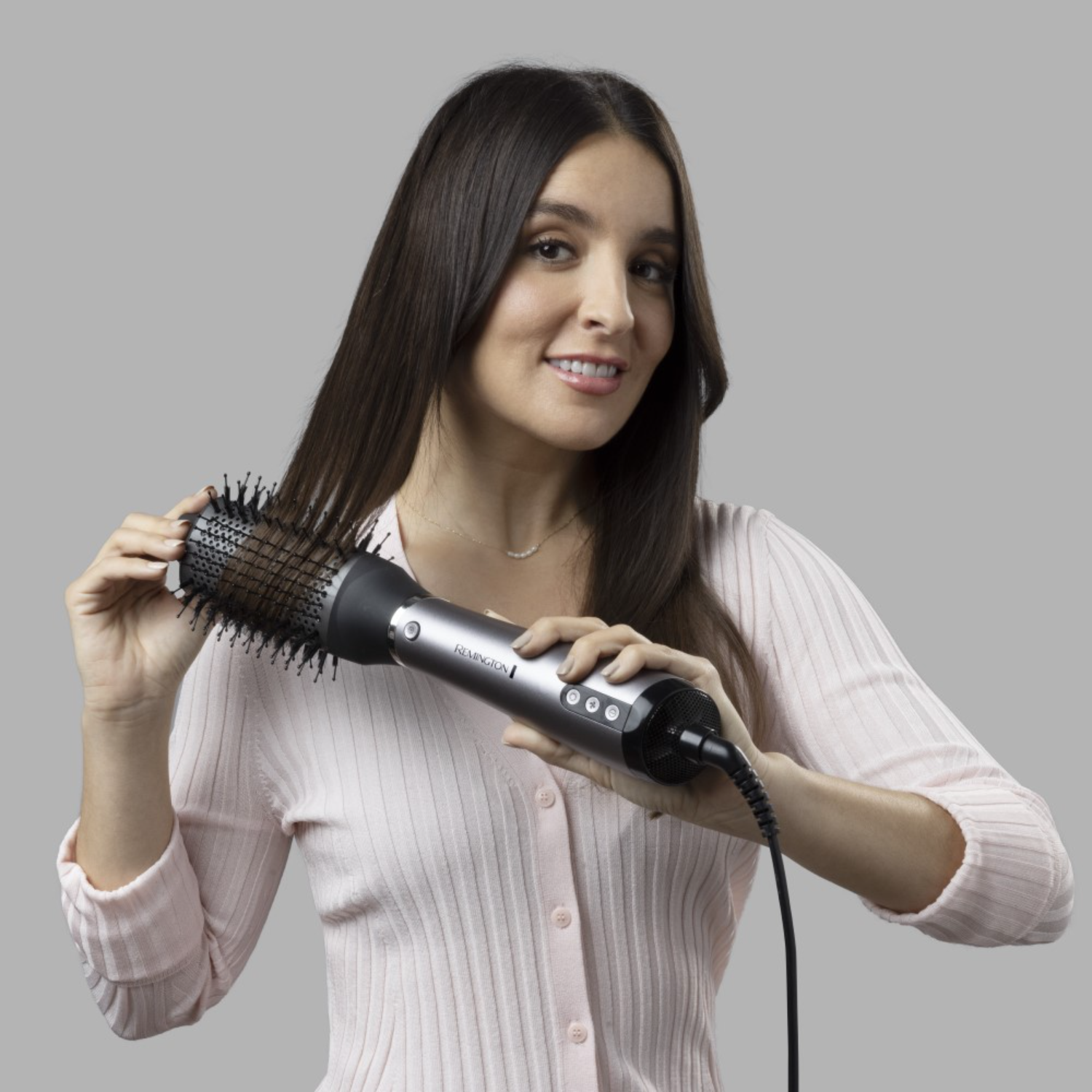 Woman styling hair with a silver and black hair styler. Light pink shirt on a gray background.