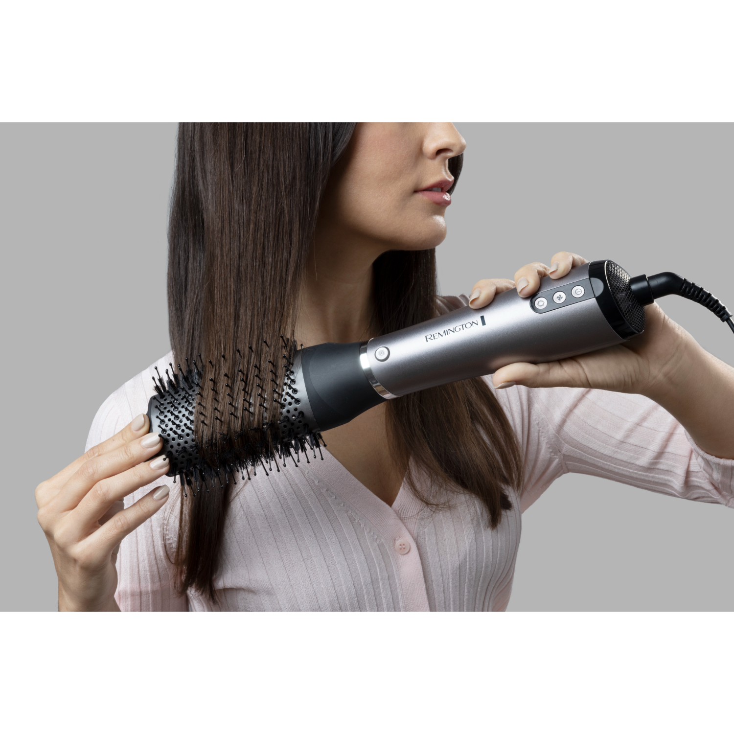 A woman holds a hair dryer brush. Her hair is being styled. The background is a light gray.