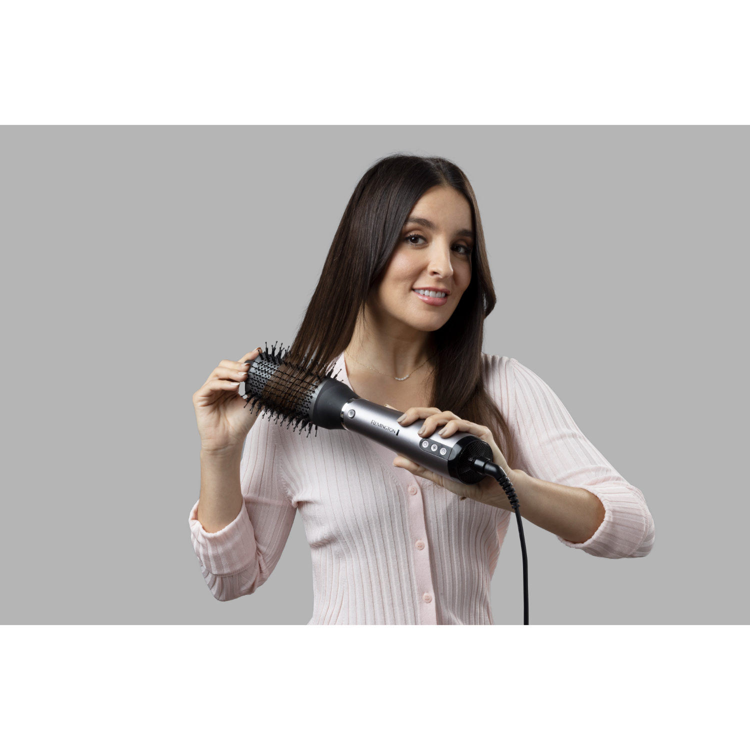 Woman styling hair with a curling brush, smiling, light pink shirt, gray background.