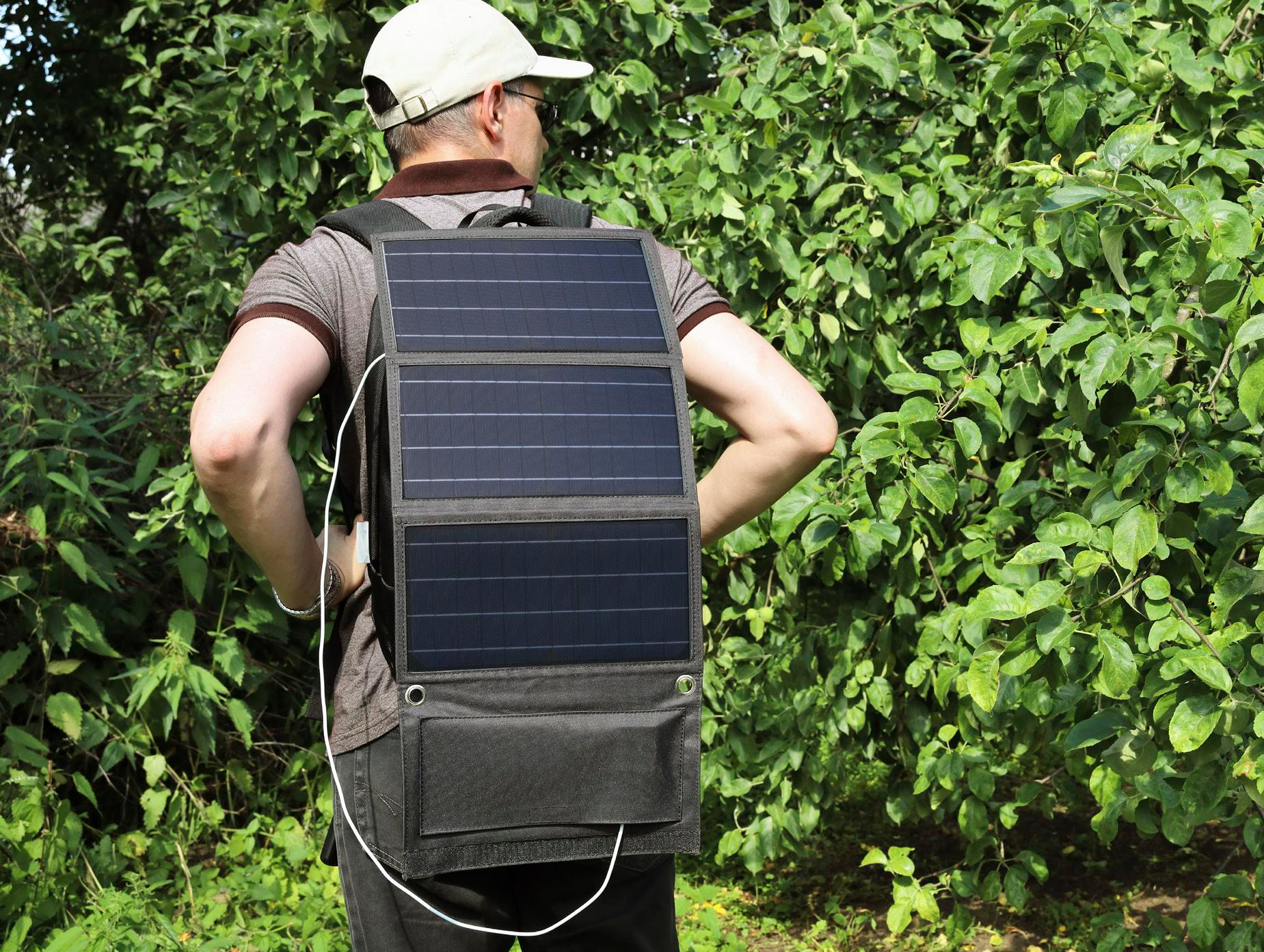 Ein Mann mit Rucksack mit Solarpanel, stehend vor einem grünen Busch, Rückansicht.