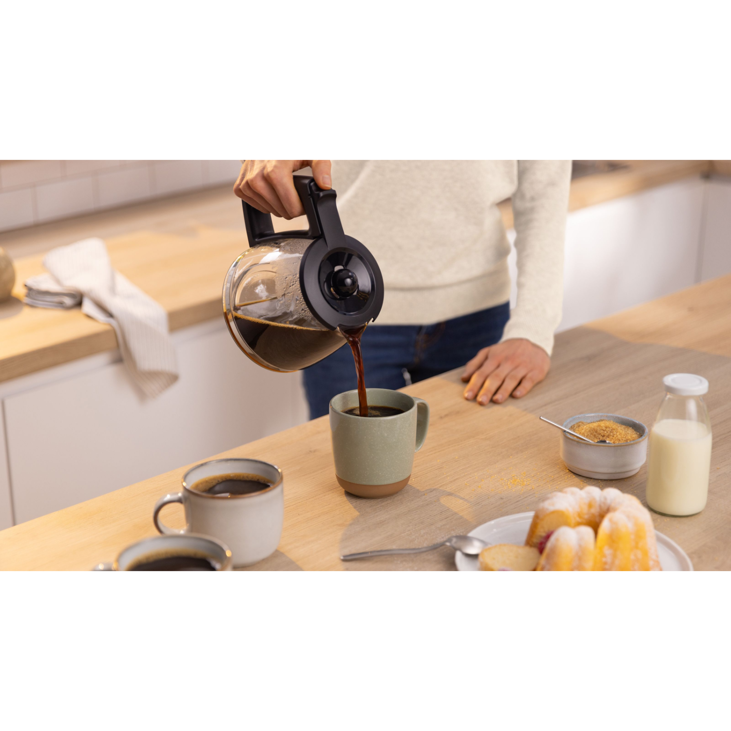 Een persoon die koffie uit een heldere kan in een groene mok giet. Een tafel met eten op de achtergrond.