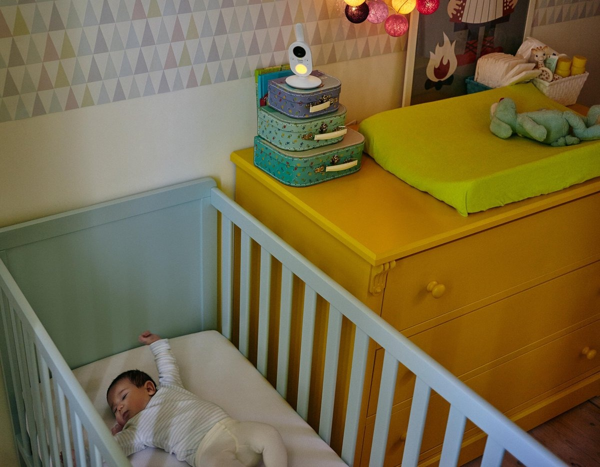 Bébé dans un berceau, bras levés, dort près d'une table à langer et d'un babyphone.