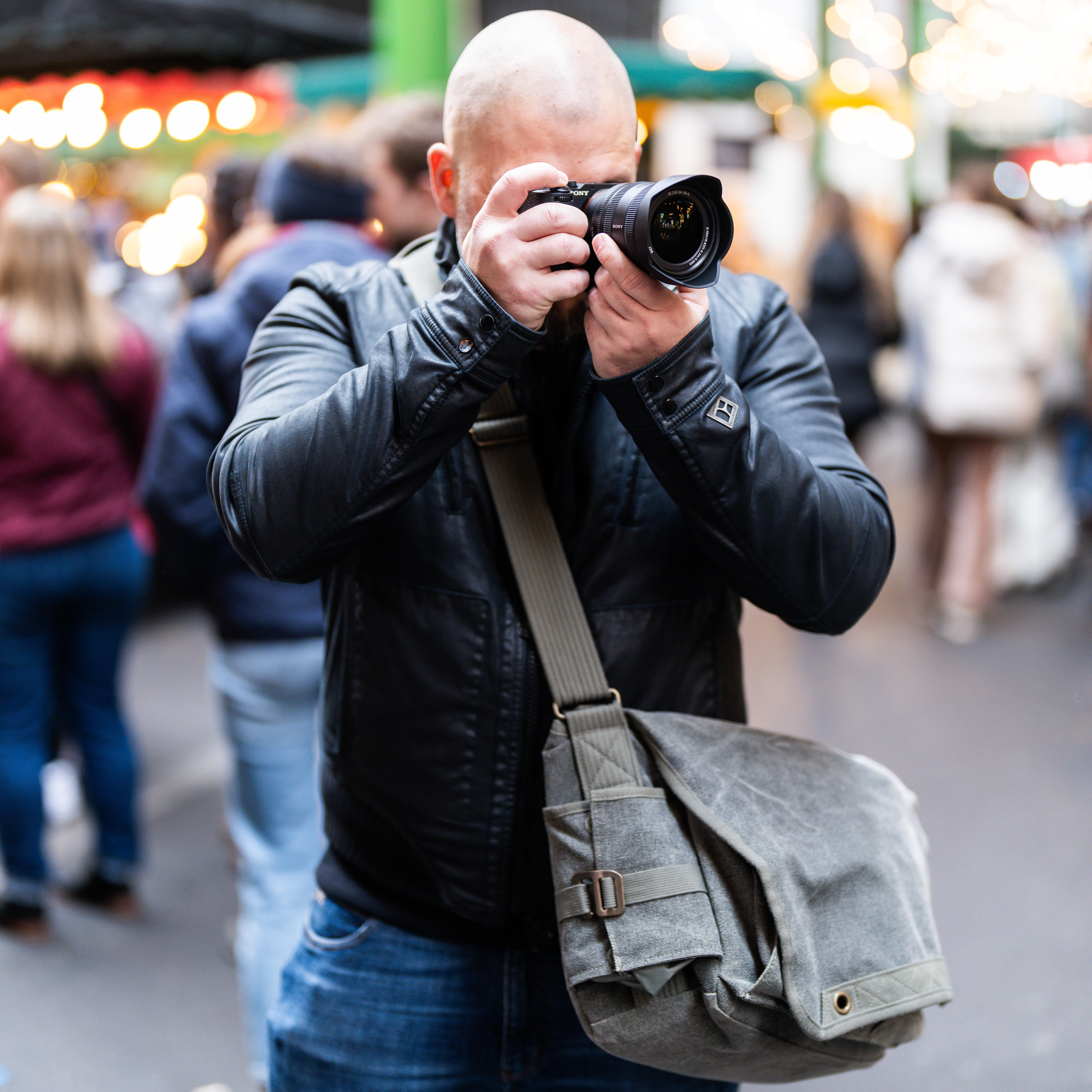 Mann, der eine schwarze Jacke trägt, hält eine Kamera und macht ein Foto. Graue Tasche auf der Schulter, verschwommene Personen im Hintergrund.