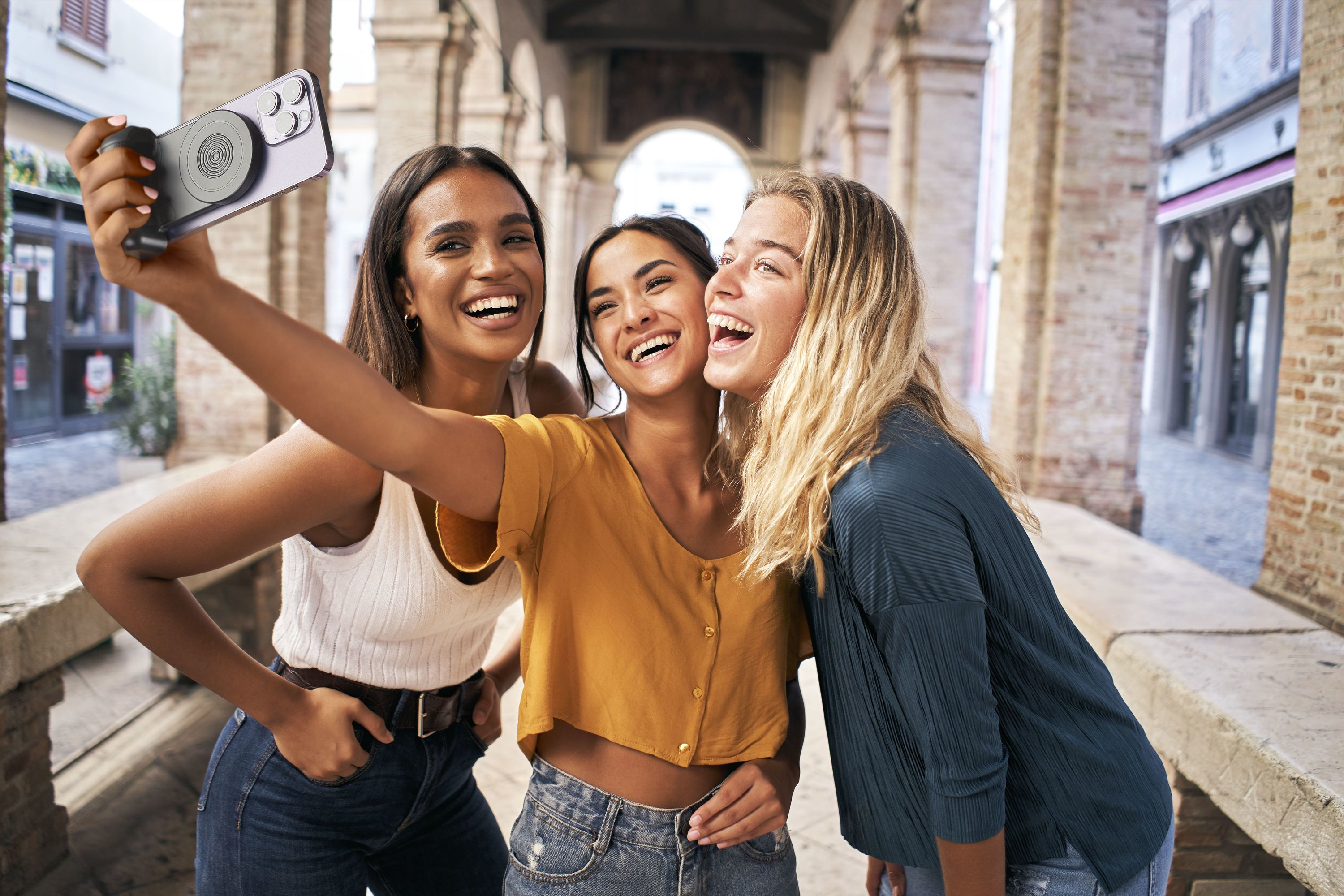 Üç kadın selfie çekiyor. Bir kemerin altında duruyorlar, gülüyorlar, arka planda bir bina var.