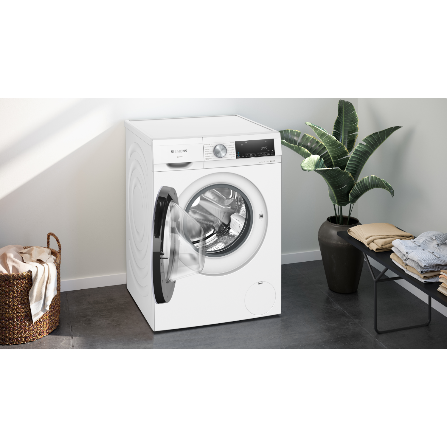 A white washing machine with the door open, next to a plant and a basket of laundry.