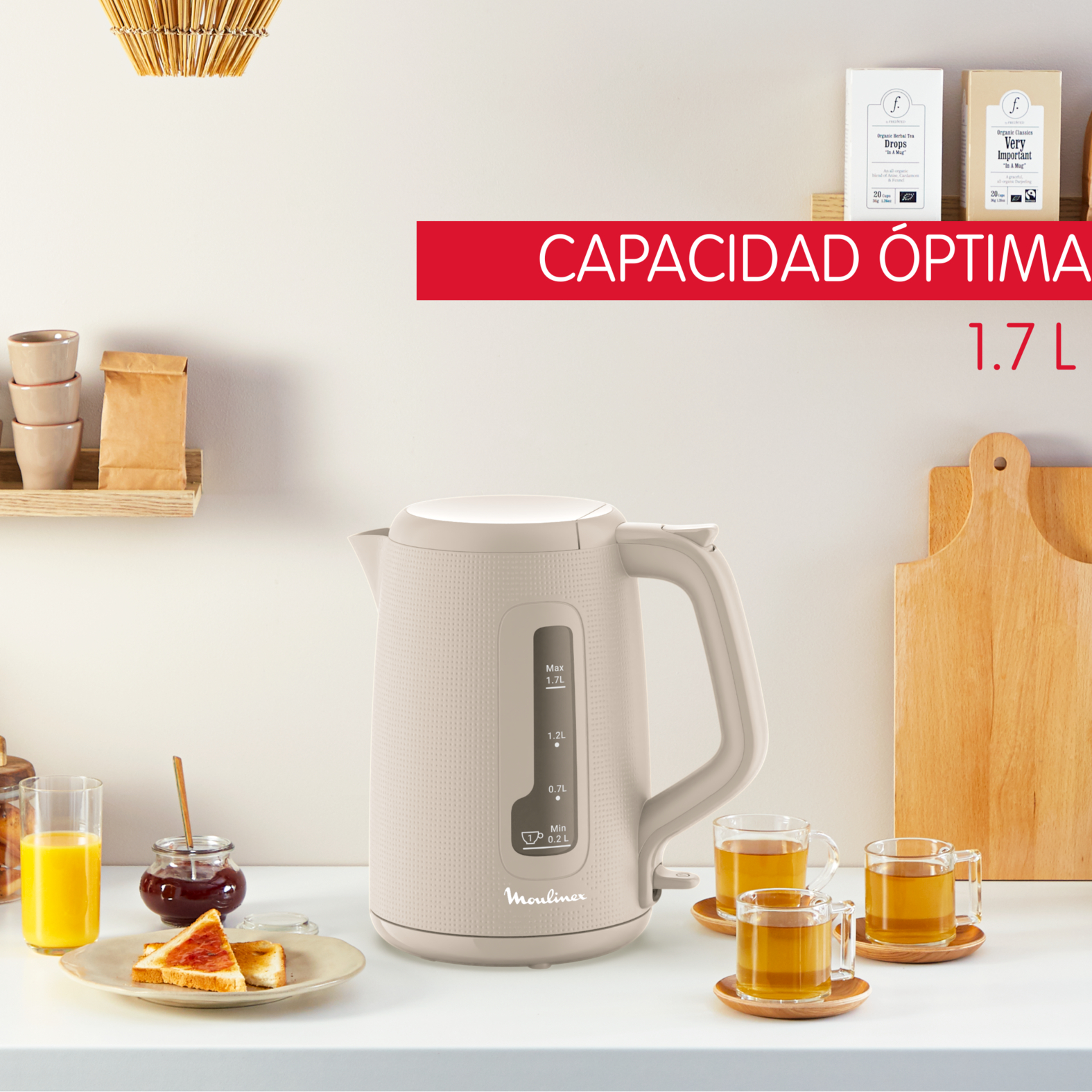 Hervidor eléctrico beige en una encimera blanca con tazas de té, pan y jugo. Texto: Capacidad 1.7 L.