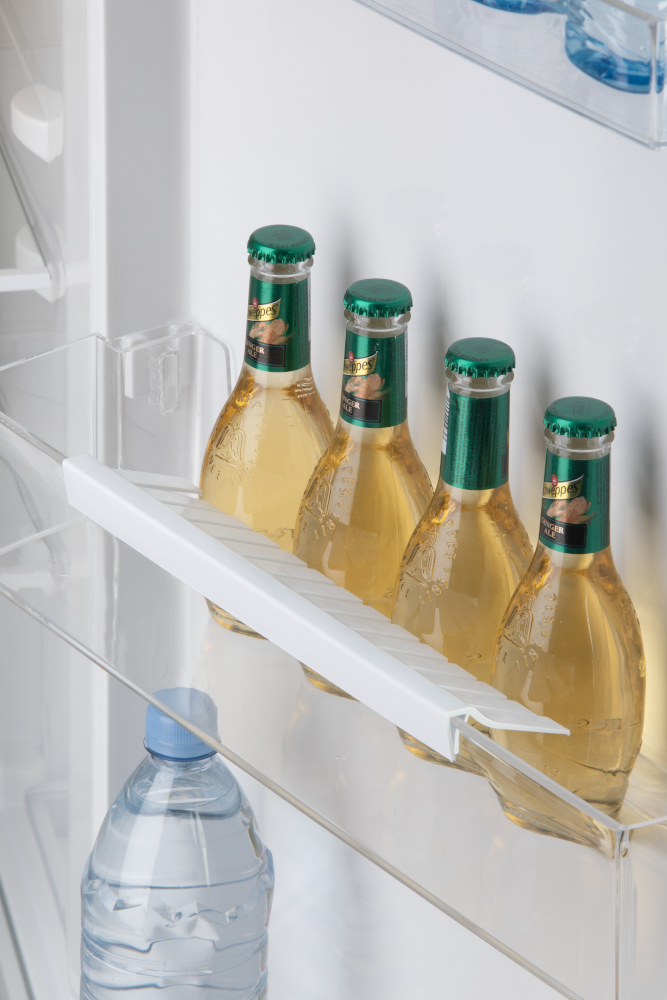 Bottles of ginger beer in fridge. Green caps, clear bottles. Refrigerator shelf and bottle of water.