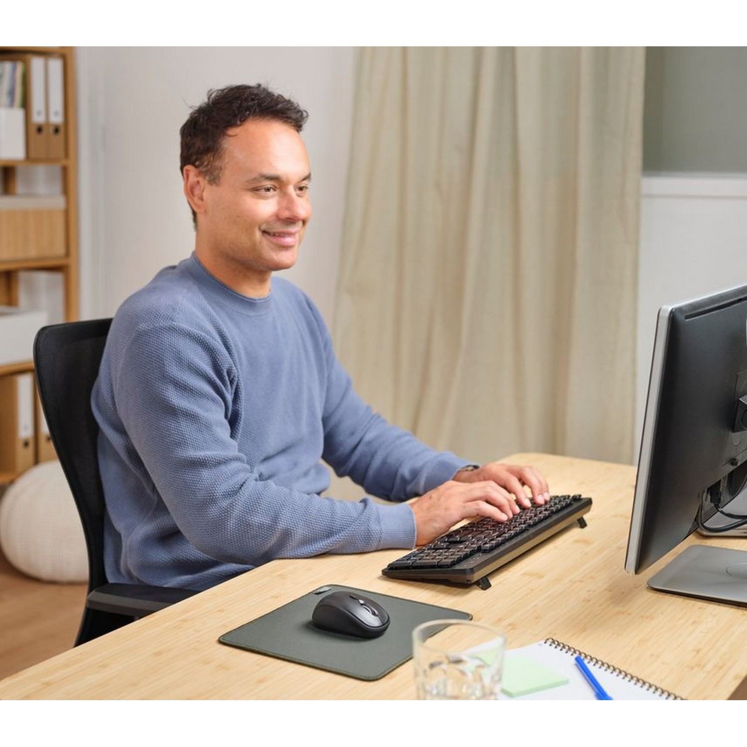 Man sitting at a desk, typing on a keyboard, looking at a monitor. Mouse and water glass are visible.