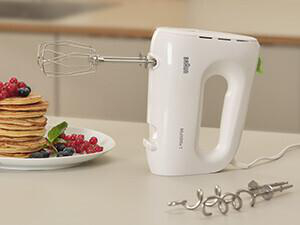 Weißer Handmixer mit Aufsätzen und Pfannkuchen, Beeren und Sahne auf einem Teller.