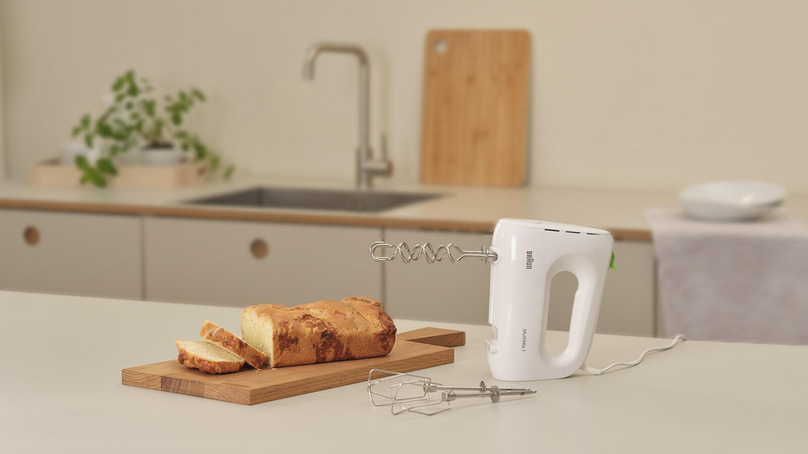 Weißer Handmixer neben geschnittenem Brot auf einem Holzbrett in einer Küche.