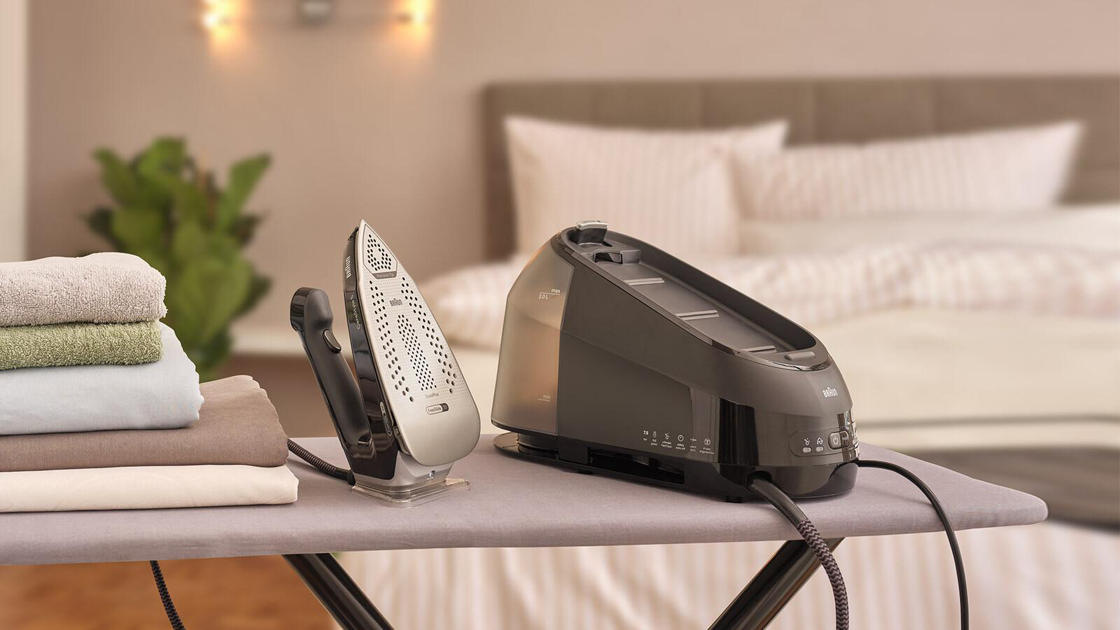Ein Braun Dampfbügeleisen und seine Basis auf einem Bügelbrett. Handtücher und ein Bett im unscharfen Hintergrund.