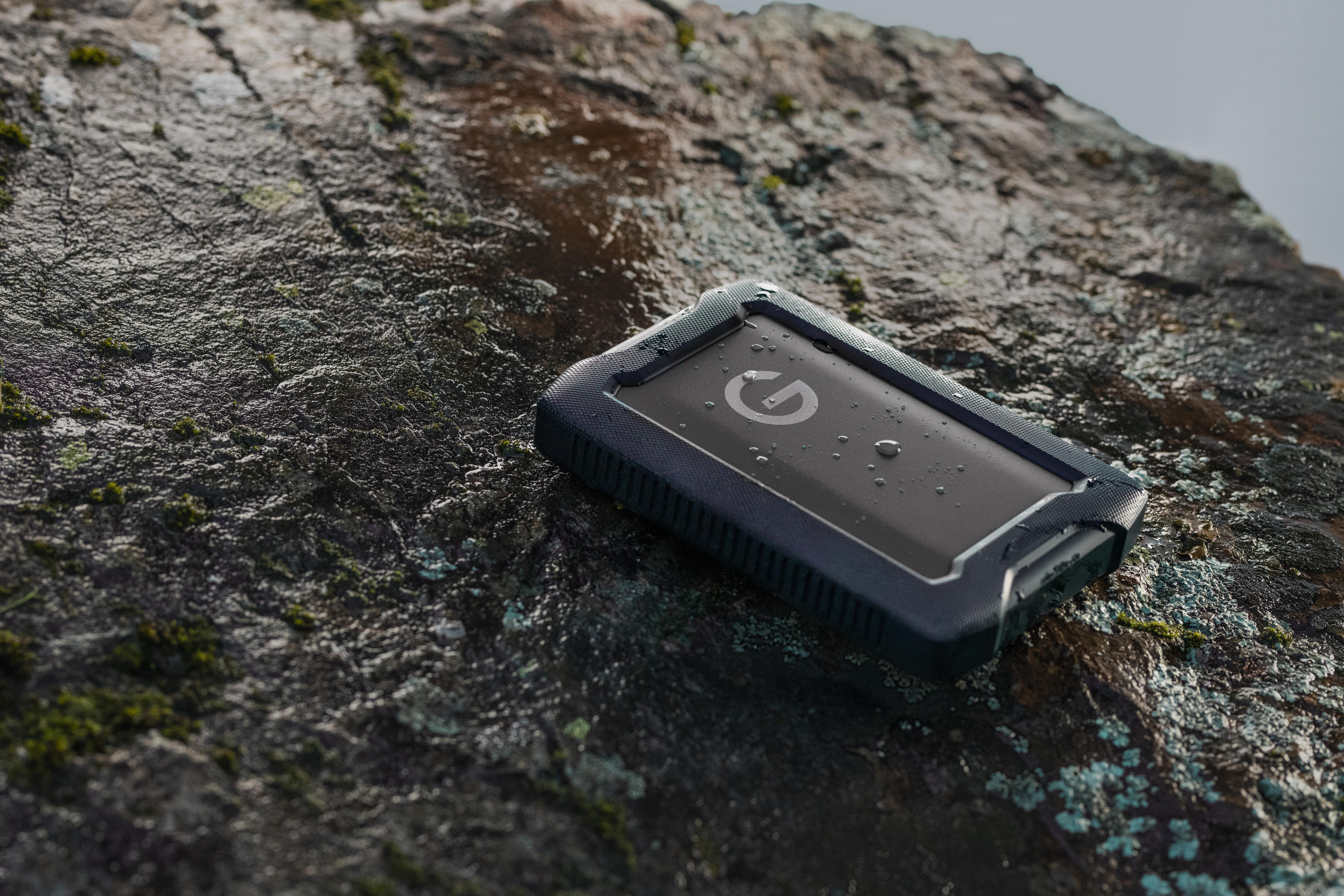 Schwarze und blaue robuste externe Festplatte auf einem Felsen, mit Wassertropfen.