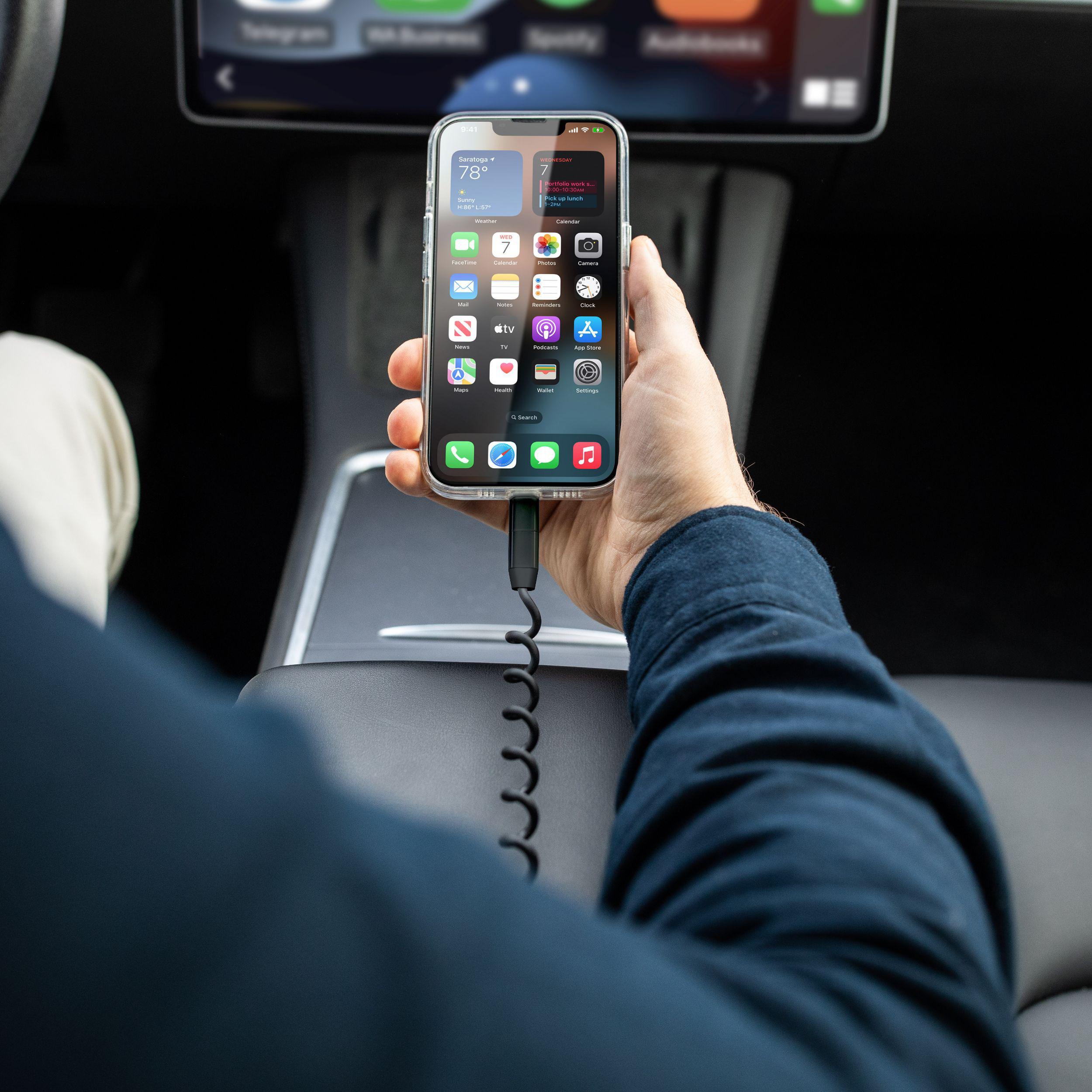 Hand, die iPhone hält, Apps sichtbar. Auto-Innenraum im Hintergrund, Ladekabel am Telefon angeschlossen.