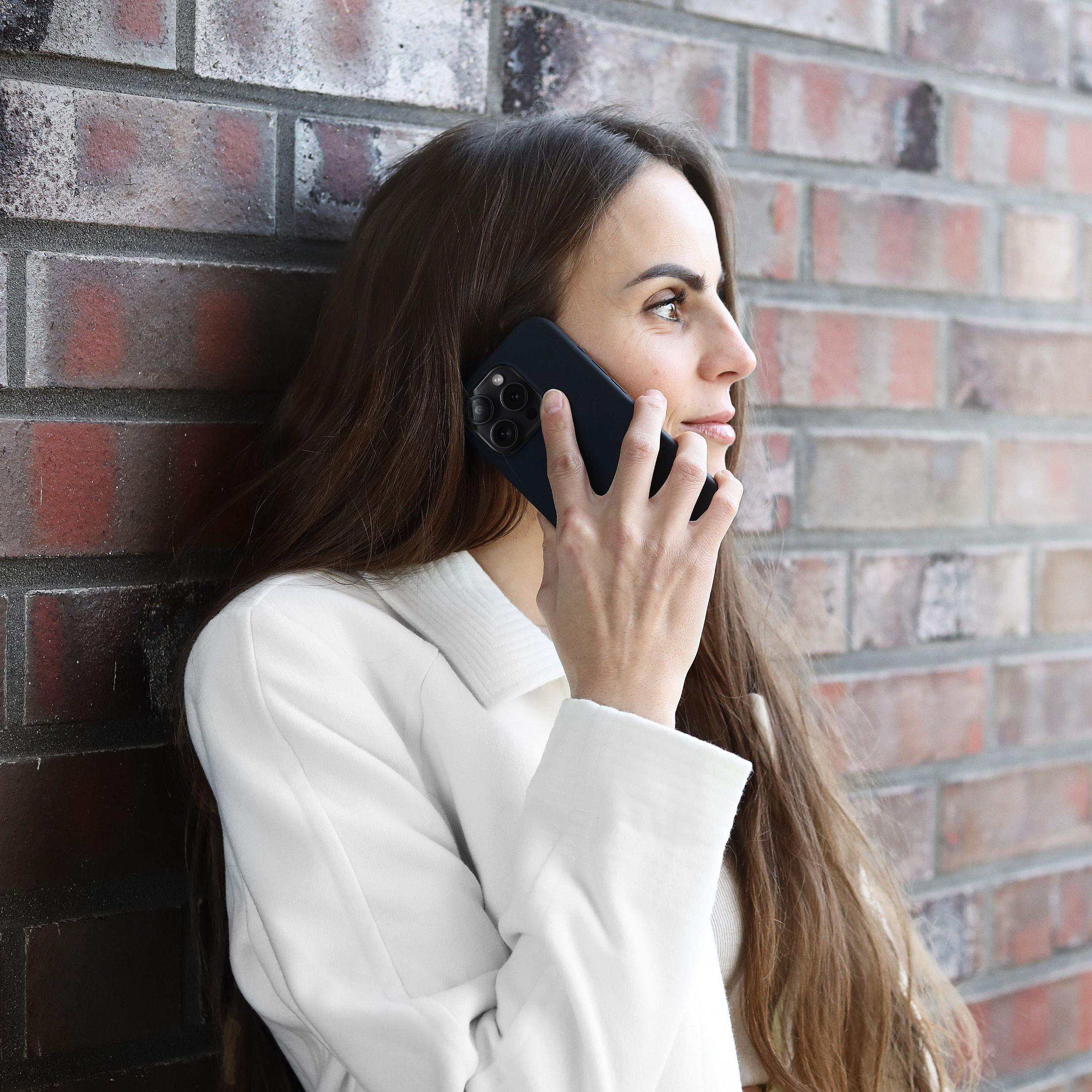 Frau in weißer Jacke lehnt an einer Backsteinmauer und telefoniert.