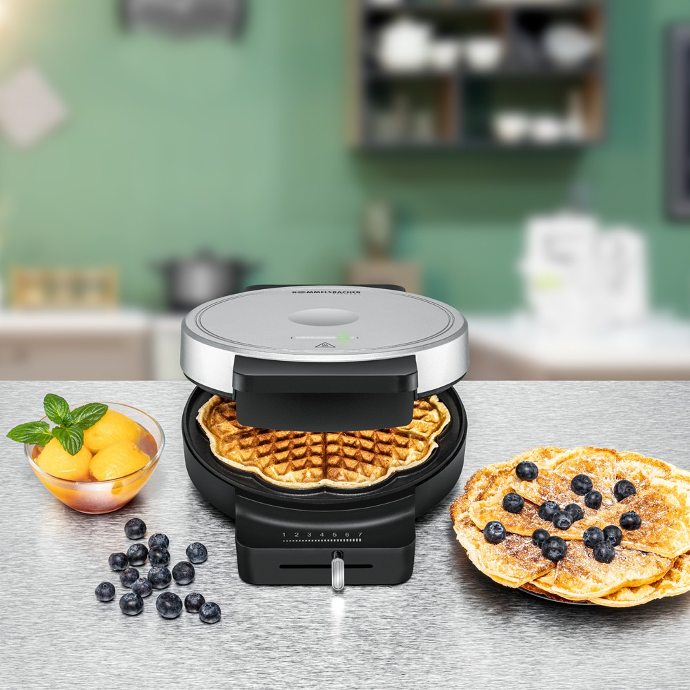 Waffeleisen mit fertiger Waffel, daneben eine Schale mit gelben Früchten, Blaubeeren und ein Teller mit Blaubeerwaffeln.