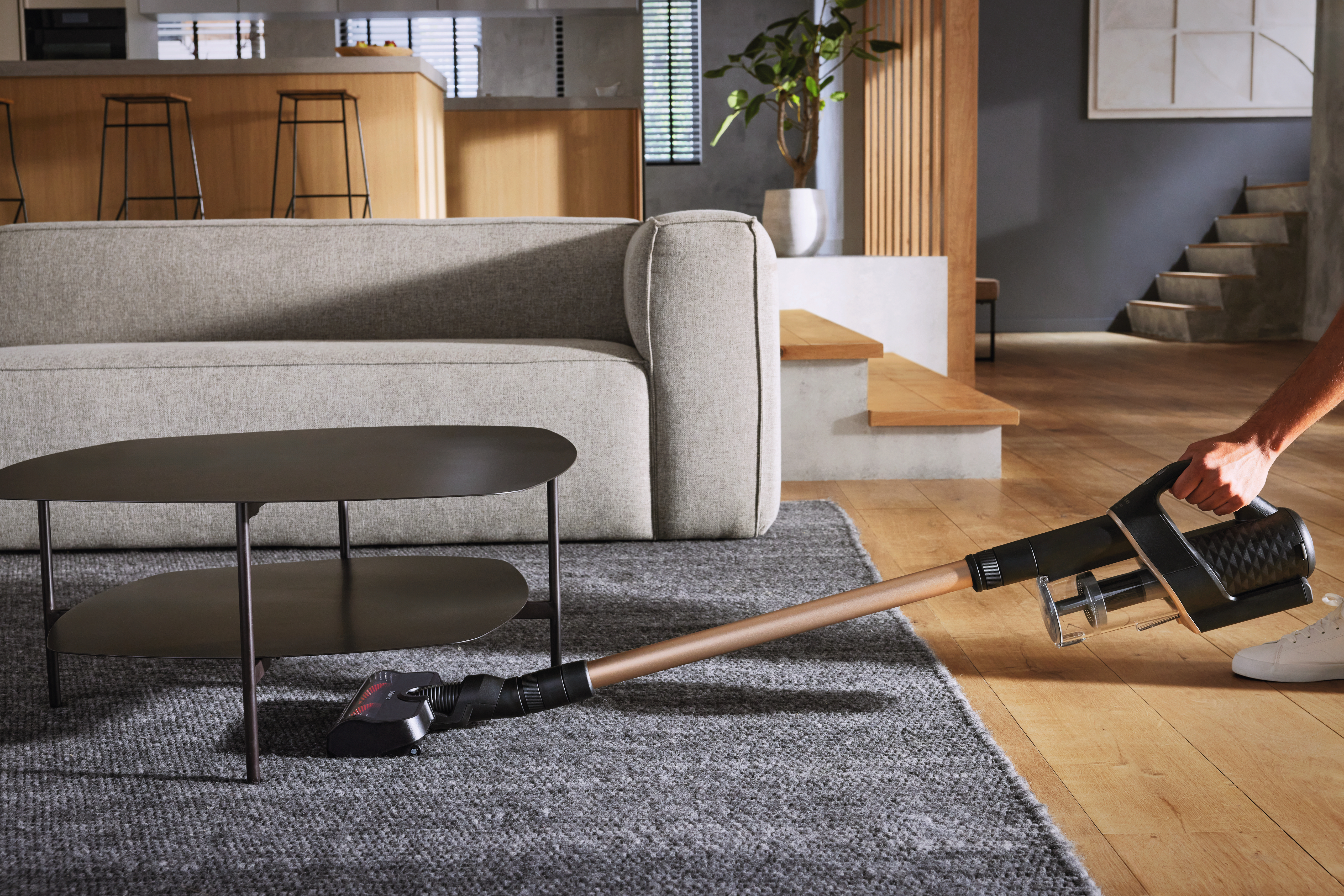 Person saugt einen Teppich mit einem schwarzen und bronzefarbenen Staubsauger. Ein Sofa und ein Tisch sind im Hintergrund.