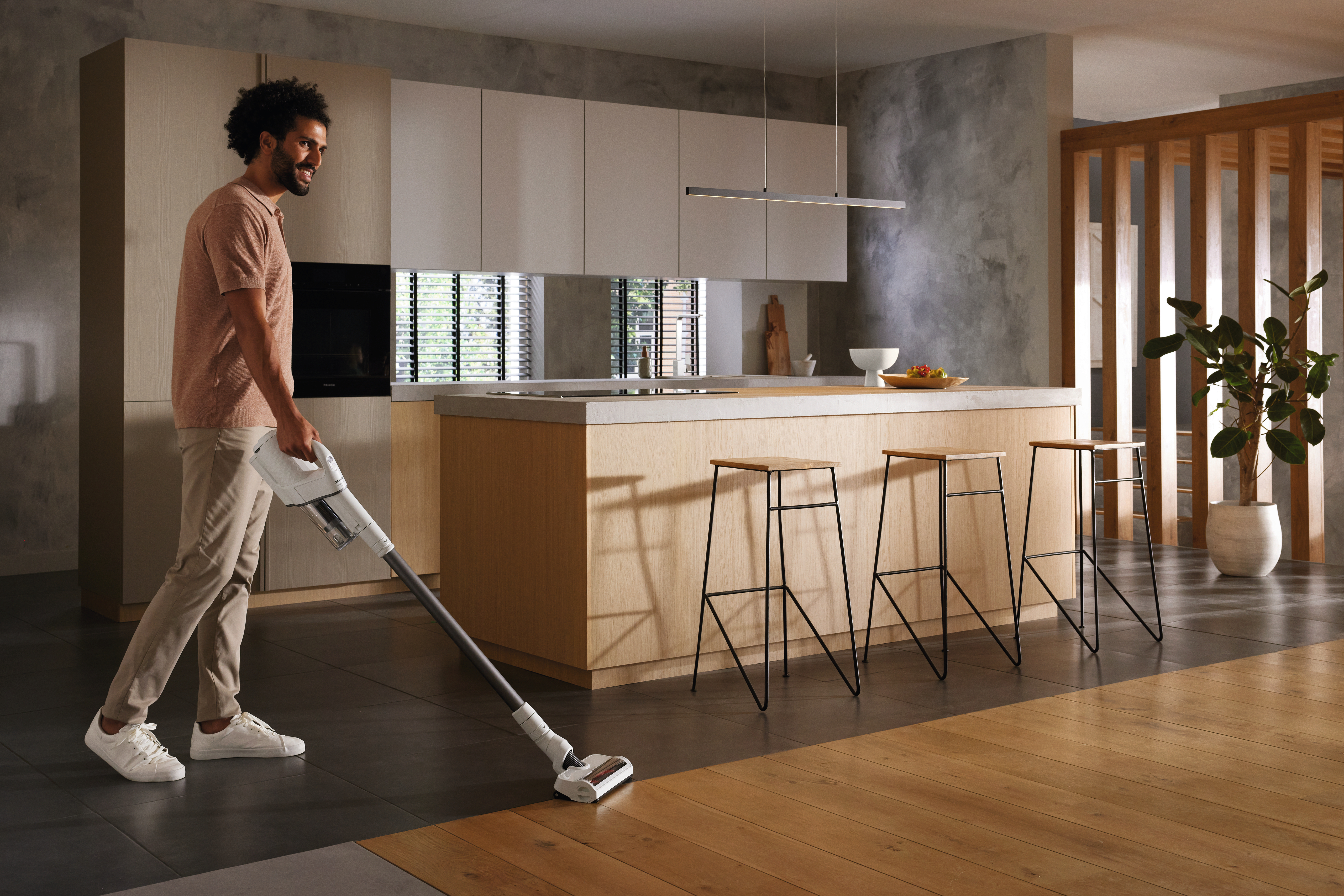 Mann, der in einer modernen Küche mit Hockern und weißen Schränken einen Holzboden saugt.