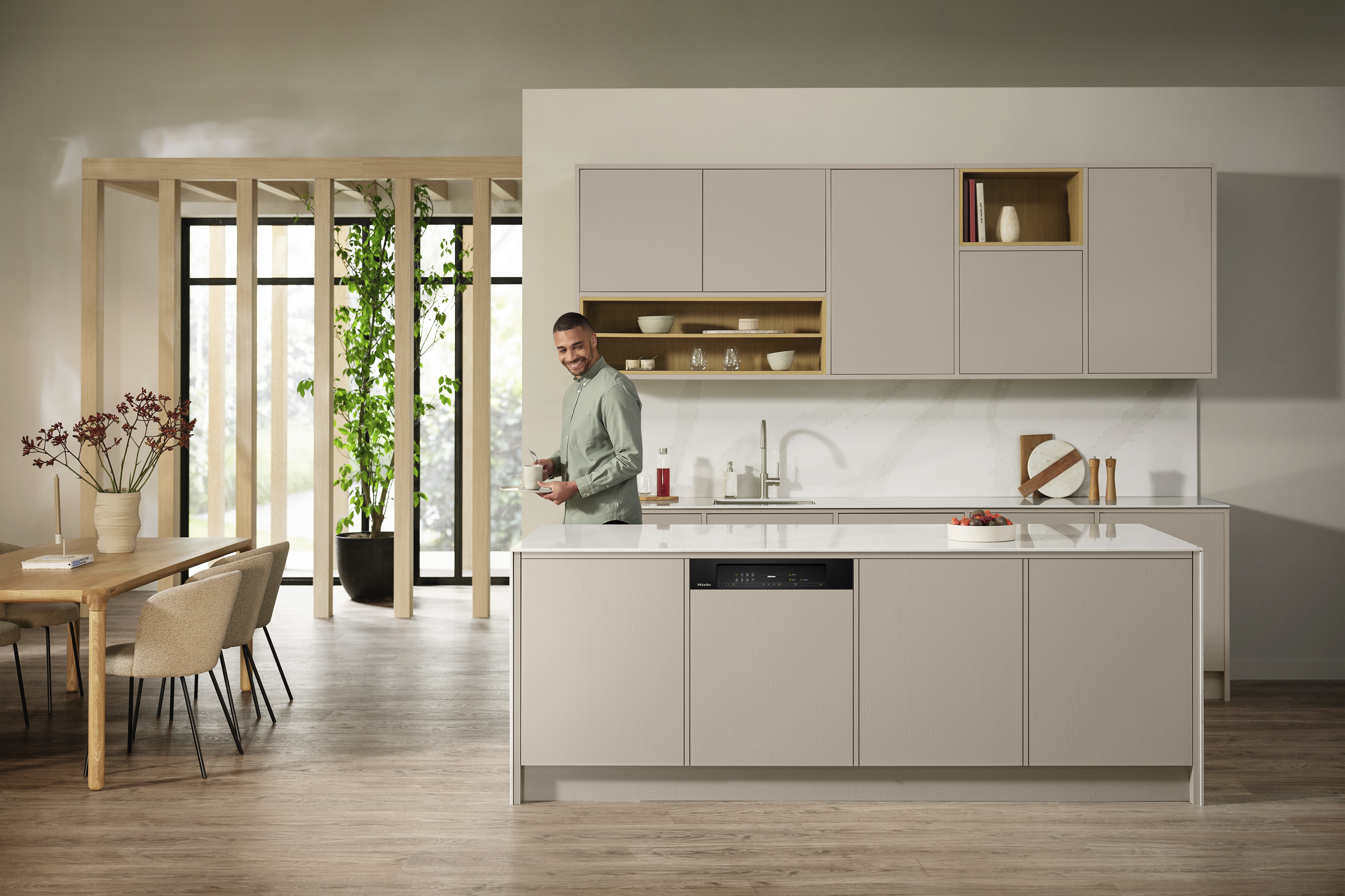 Ein Mann in einer modernen Küche mit Geschirrspüler. Beige Schränke und Insel, Holztisch.