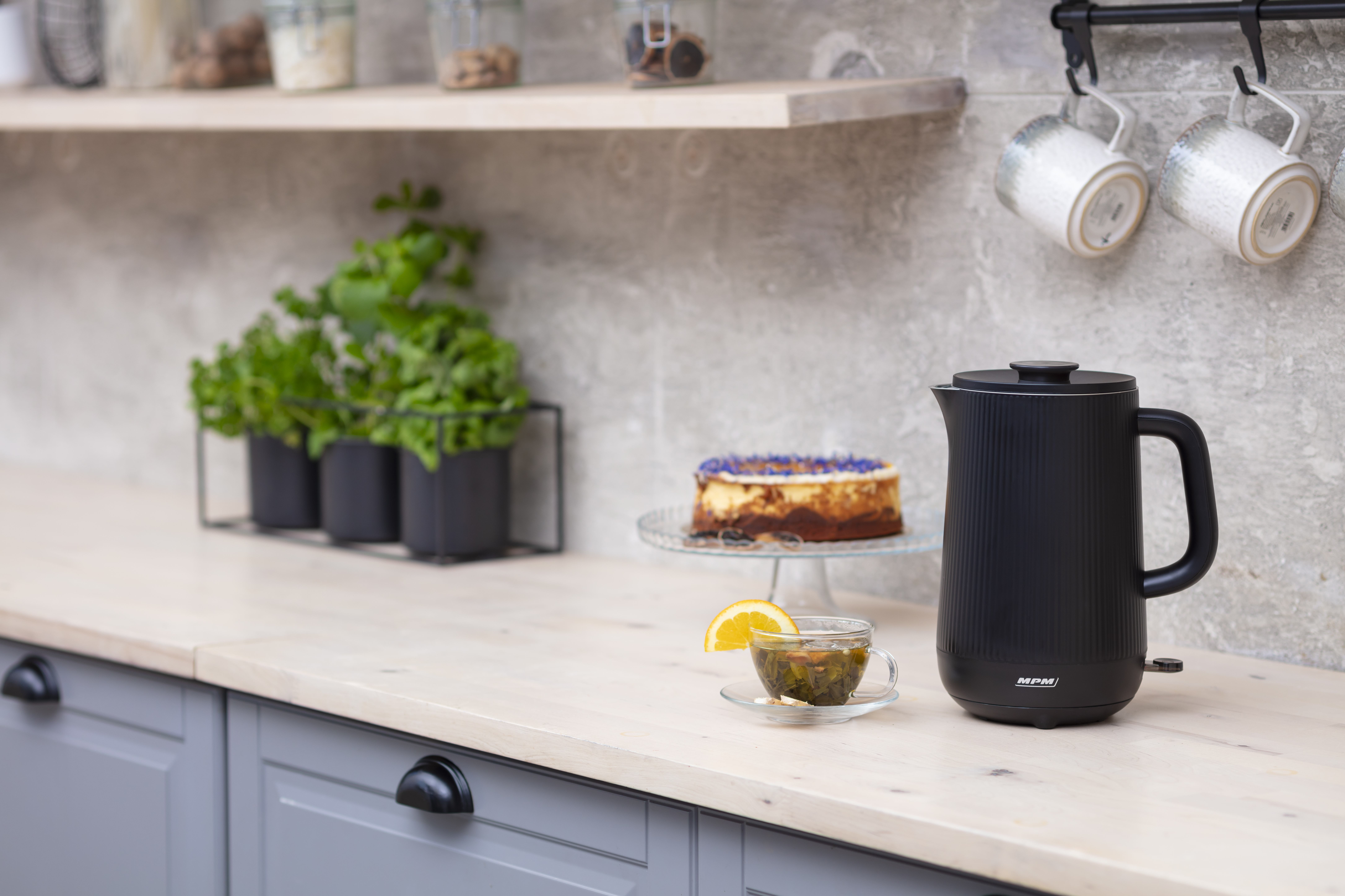 Czarny czajnik elektryczny i filiżanka herbaty na drewnianym blacie. Ciasto i rośliny w tle.