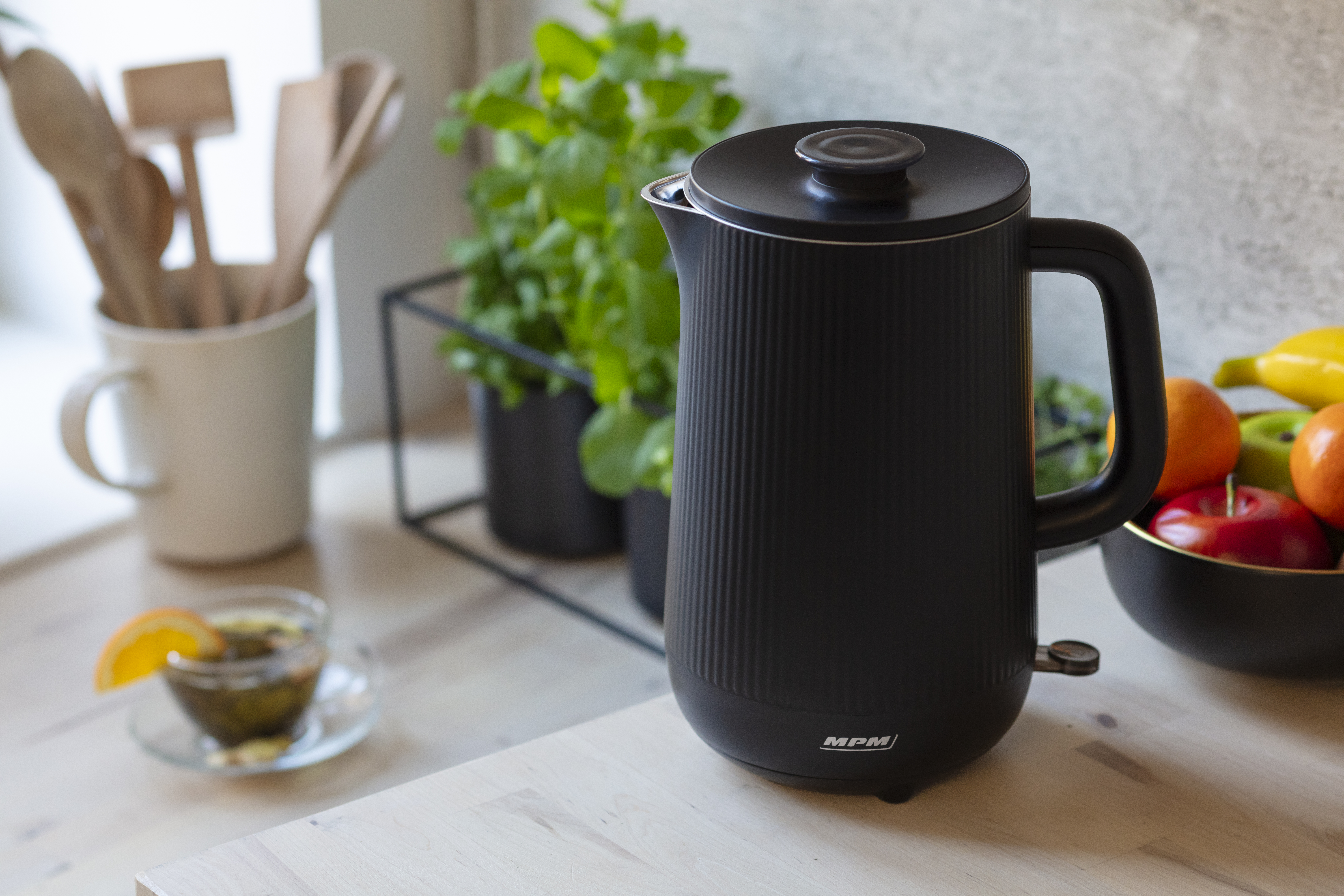 Czarny czajnik elektryczny na drewnianym blacie. Roślina i owoce po prawej.