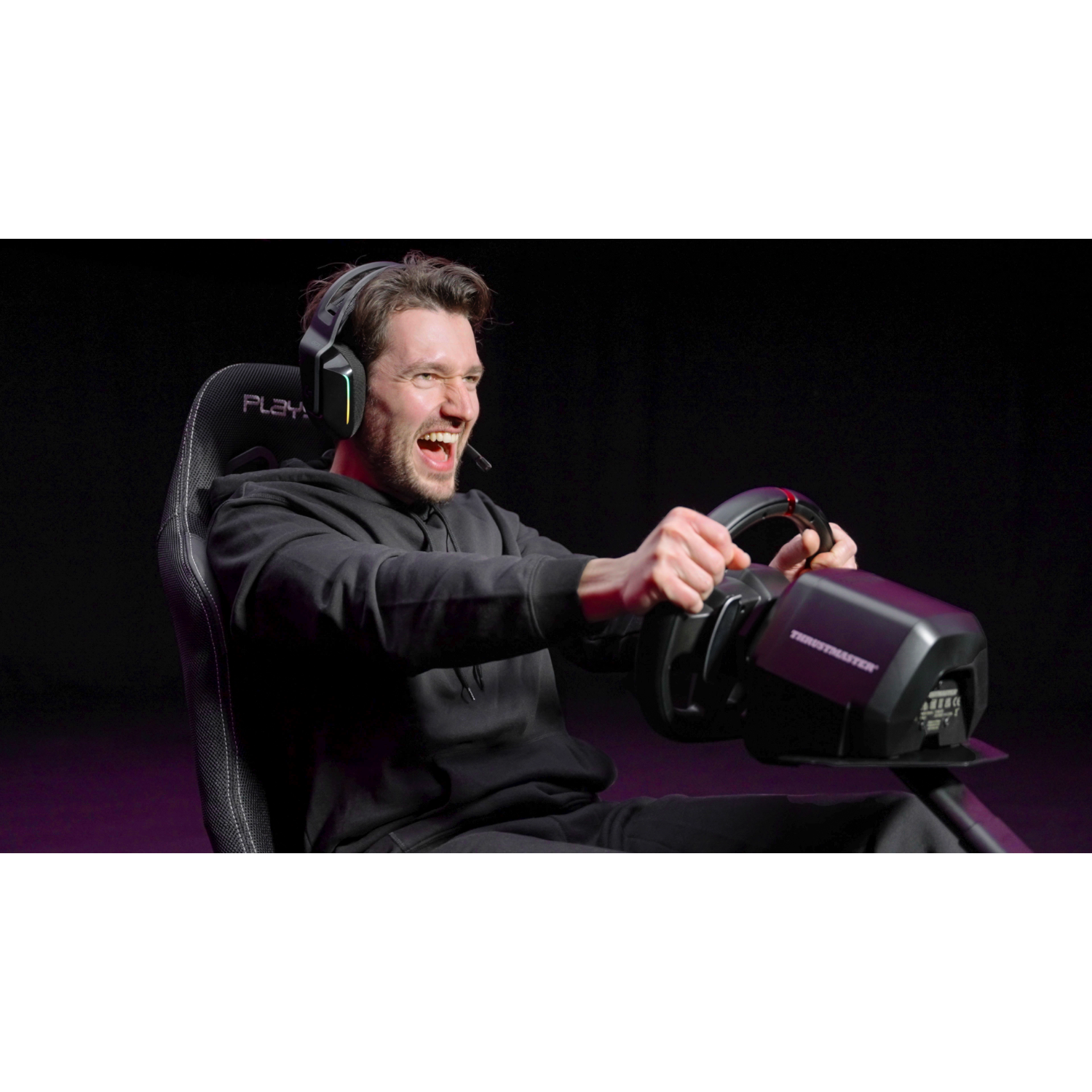 Man in headphones and racing wheel, eyes wide, mouth open. He's gaming, dark background, lit with purple and green.