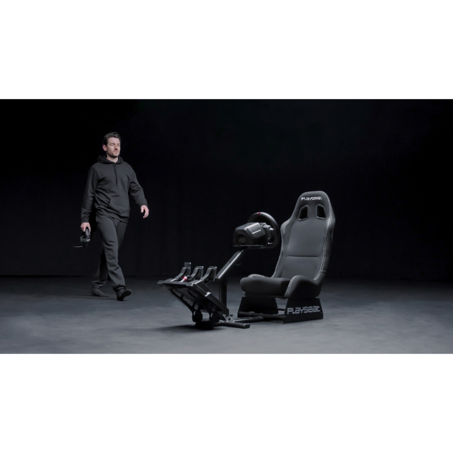 Man walking toward racing simulator seat. Black seat, steering wheel, and pedals. Dark background.